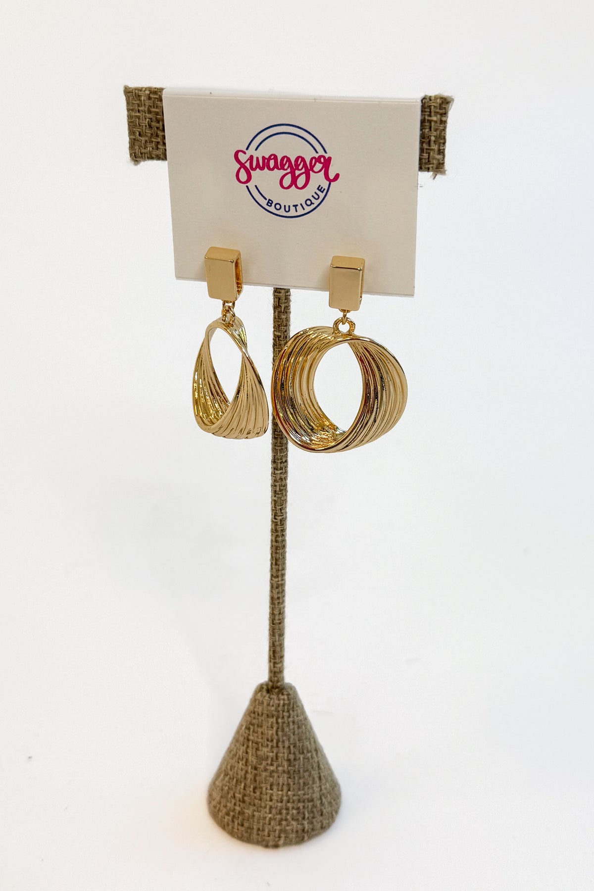 A pair of gold-toned statement earrings displayed on a burlap-covered stand with a white card labeled &