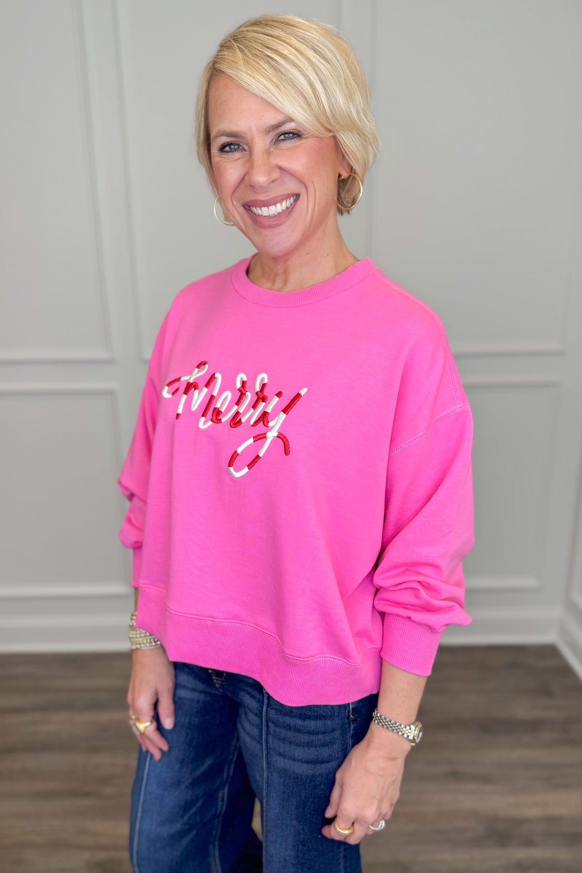 A smiling blonde woman wearing a bright pink crewneck sweatshirt with &