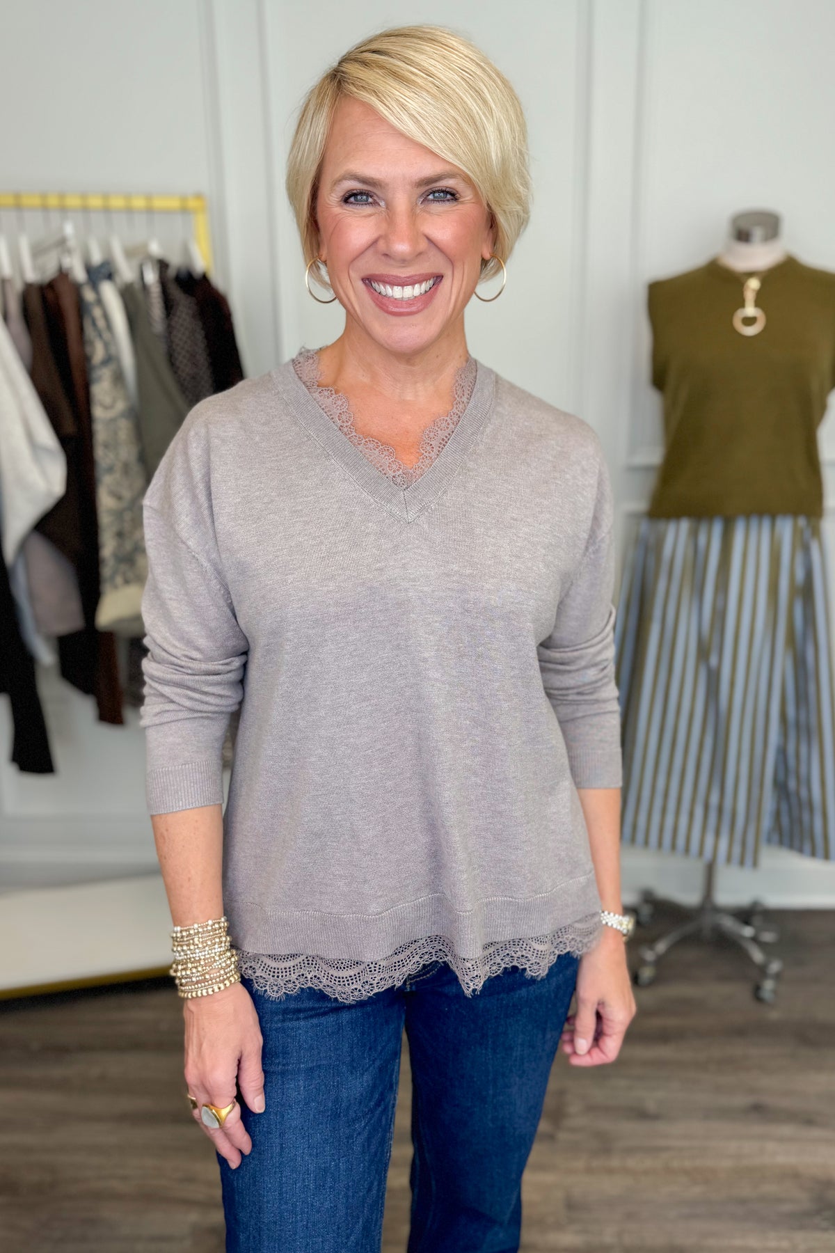 Front view of woman wearing Swagger Boutique’s Mocha Lace Lined V-Neck Sweater with jeans with clothing hanging in the background.