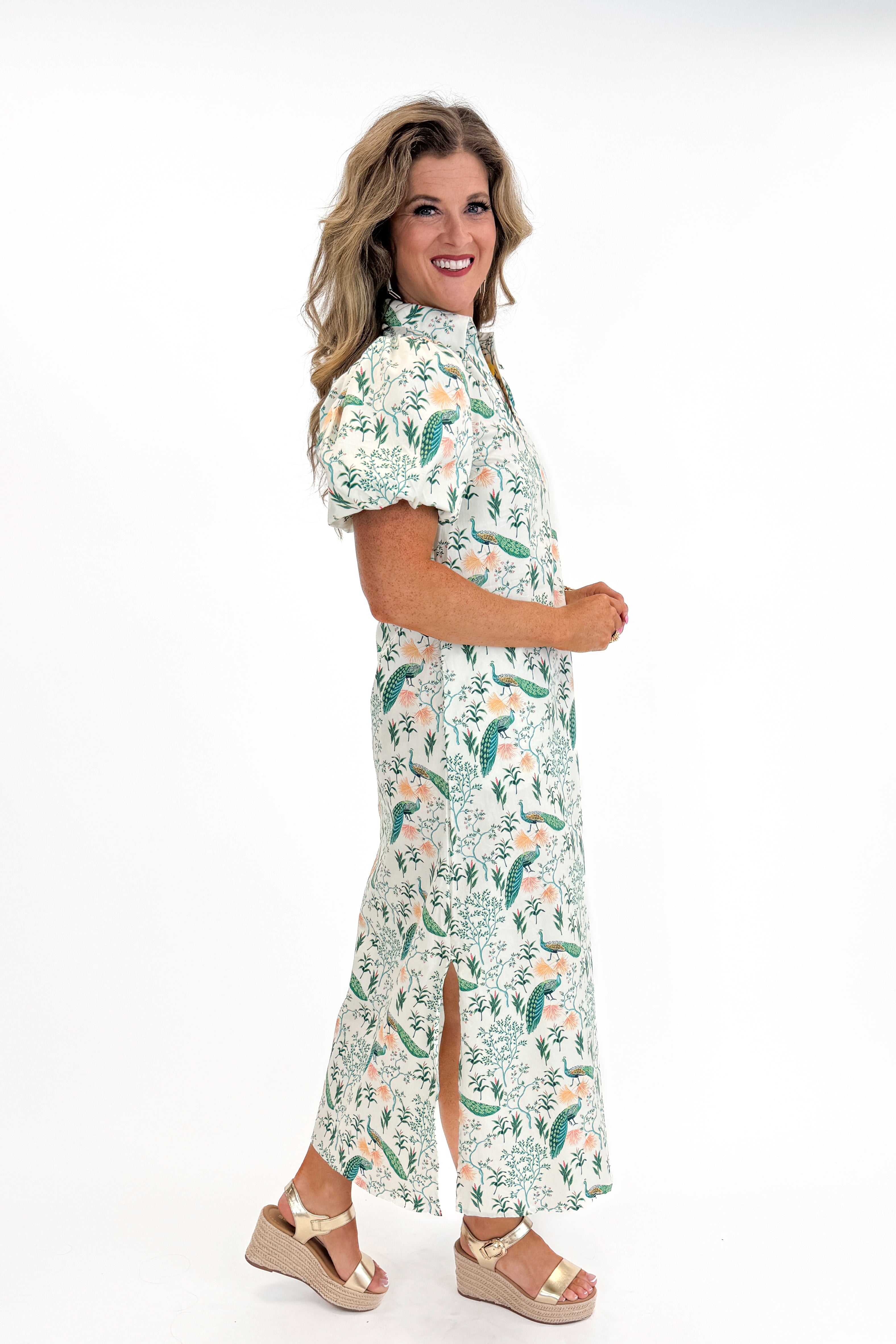 Side view of woman wearing Swagger Boutiqueβs Multi Peacock Print Maxi Dress with sandals against white studio background.
