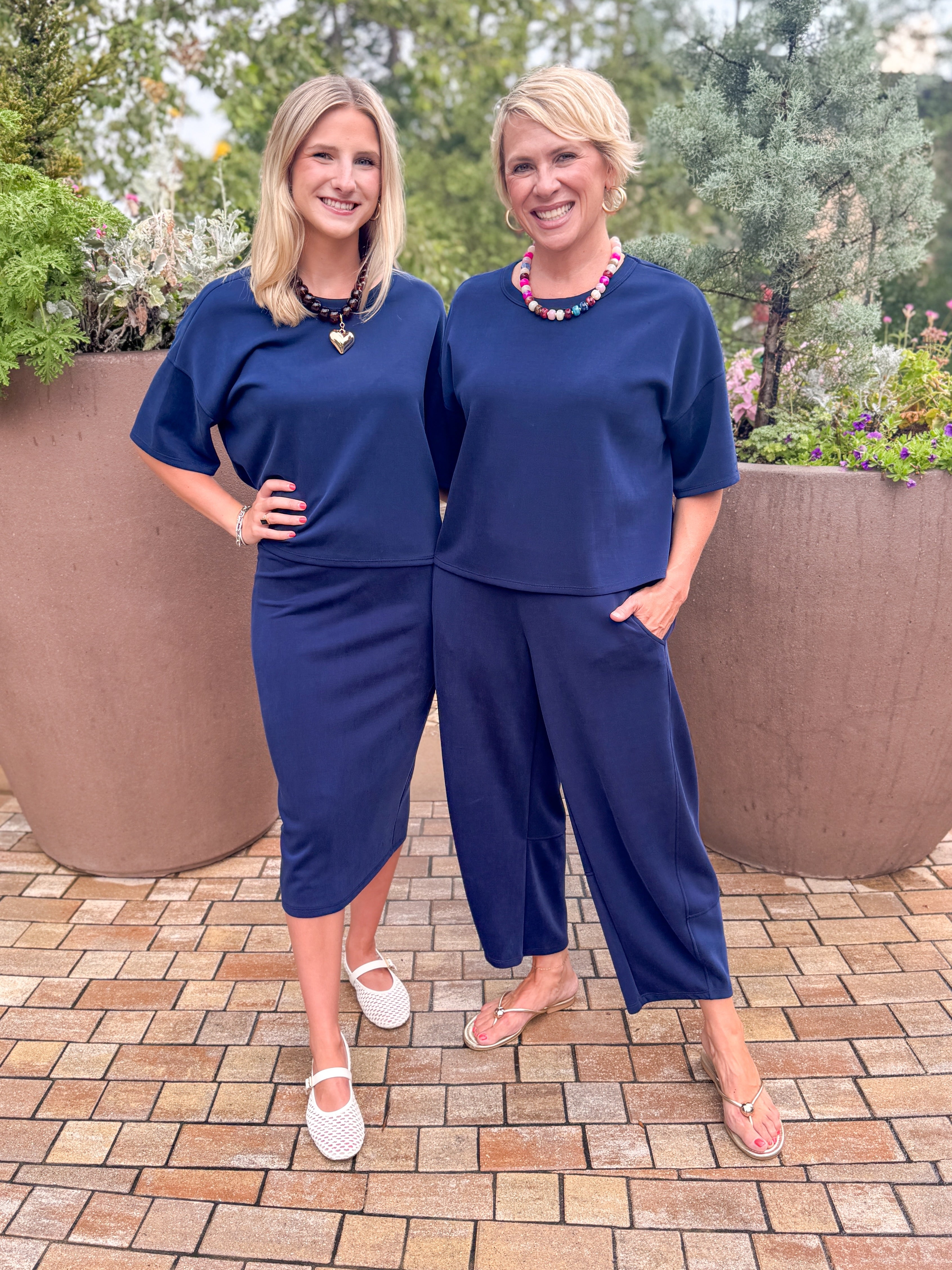Front view of woman wearing Swagger Boutiqueβs Navy Barrel Lounge Pants with Navy Round Neck Short Sleeve Top and woman wearing same top and matching skirt standing outside at a park.