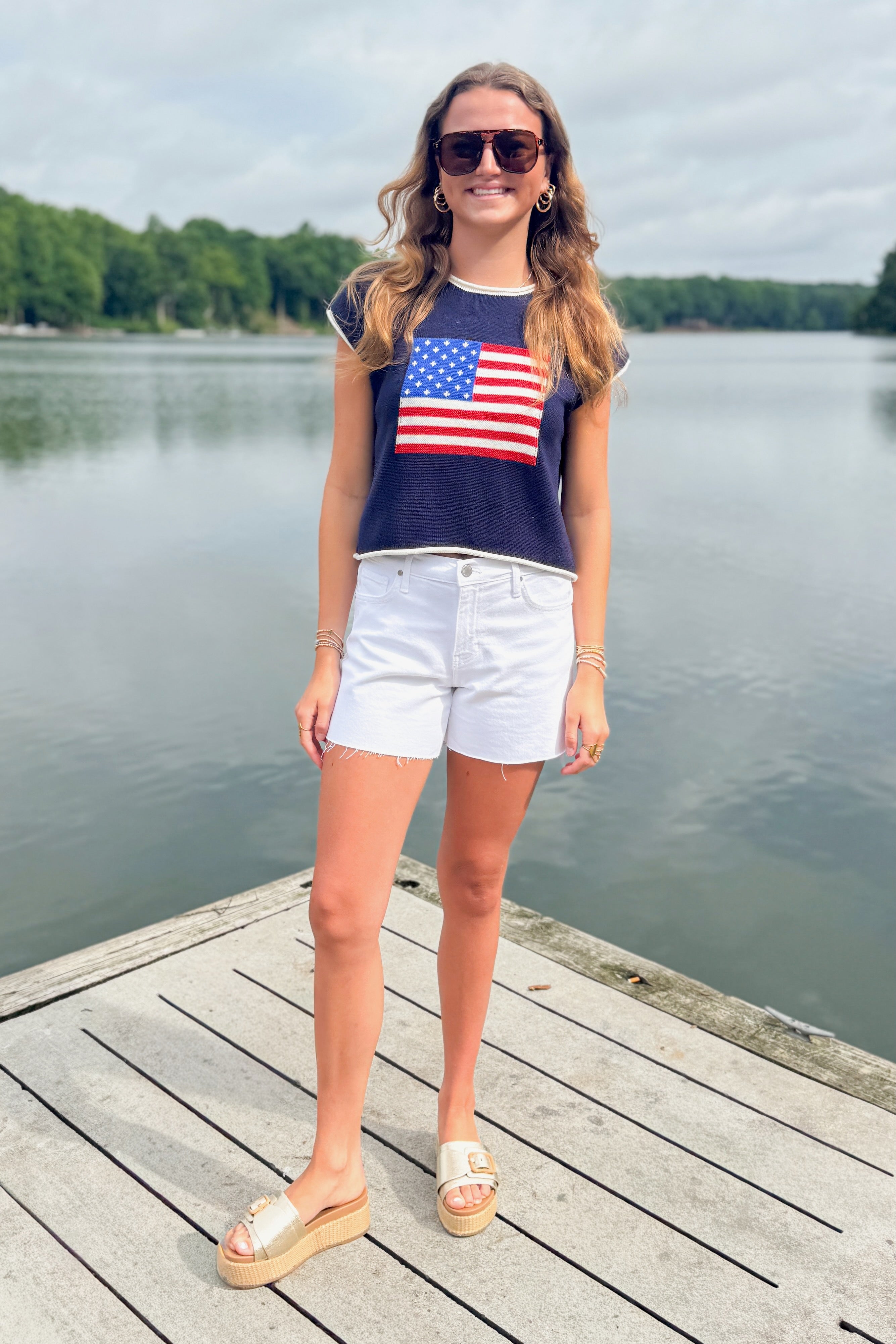 Front view of woman wearing Swagger Boutiqueโs Right On Cue Navy Flag Short Sleeve Sweater with jean shorts at the lake.