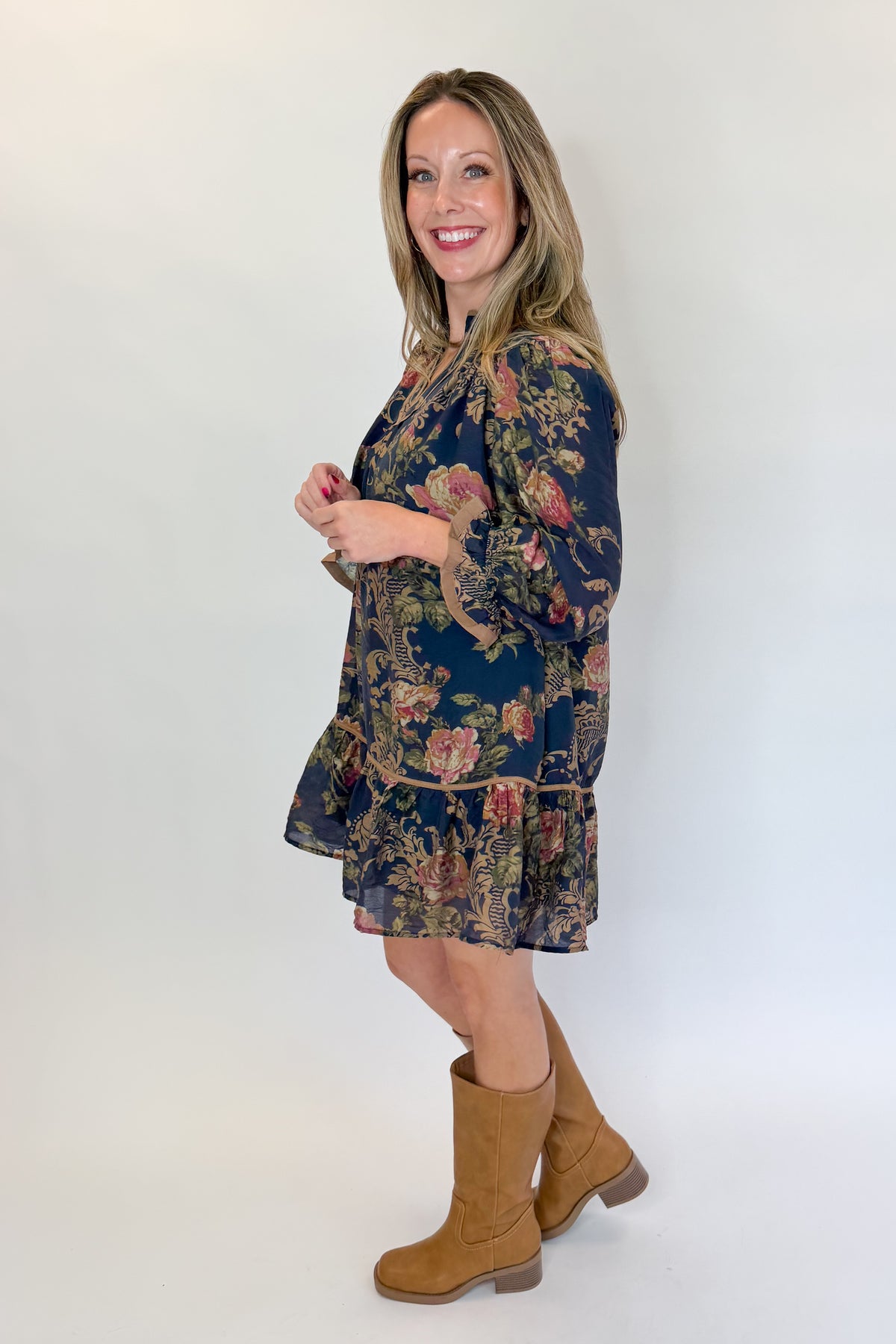 Side view of woman wearing Swagger Boutique’s Navy Floral Print Mini Dress with boots against white studio background.