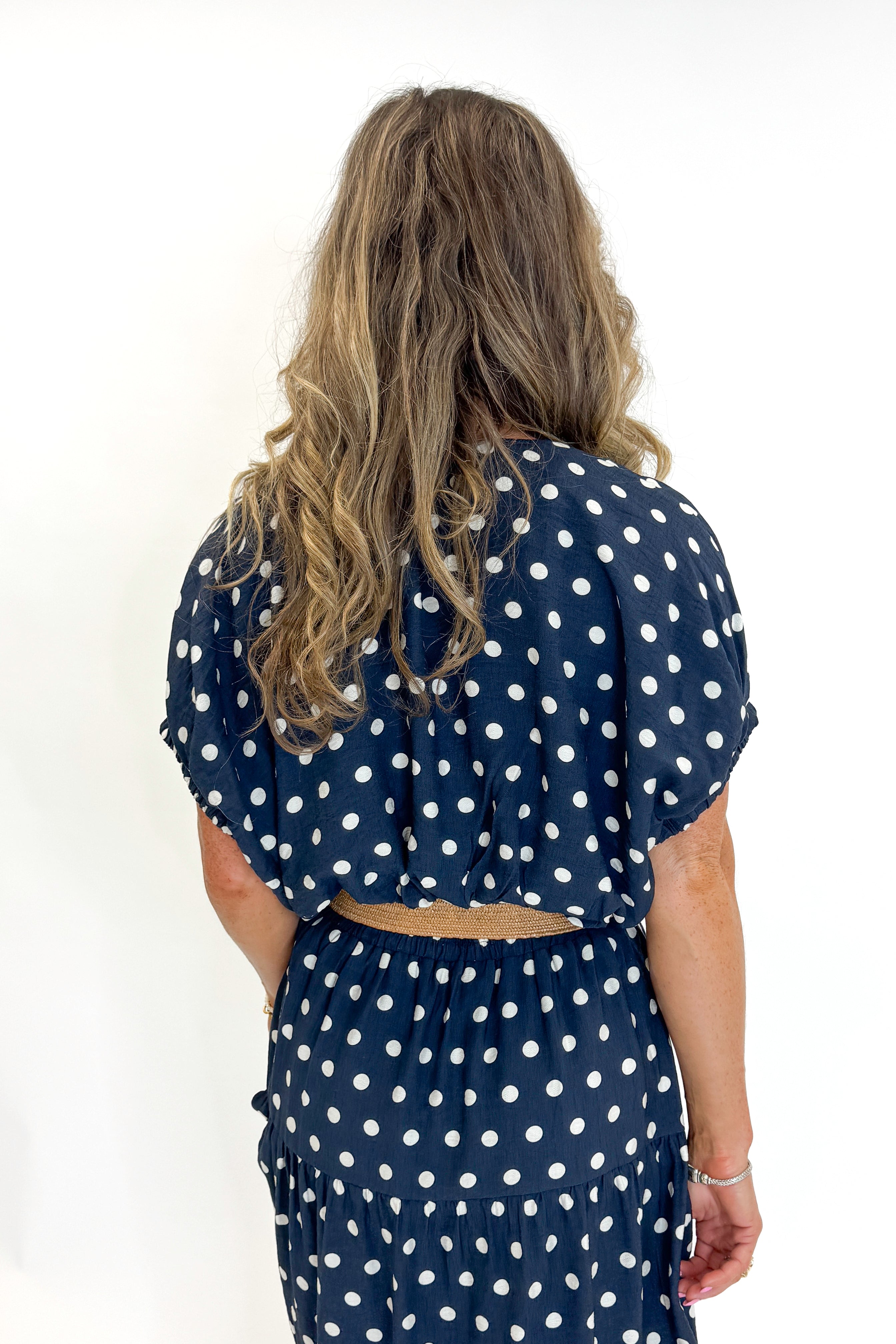 Back view of woman wearing Swagger Boutiqueβs Navy Polka Dot Bubble Hem Top by Entro with matching Navy Polka Dot Tiered Skirt by Entro against white studio background.