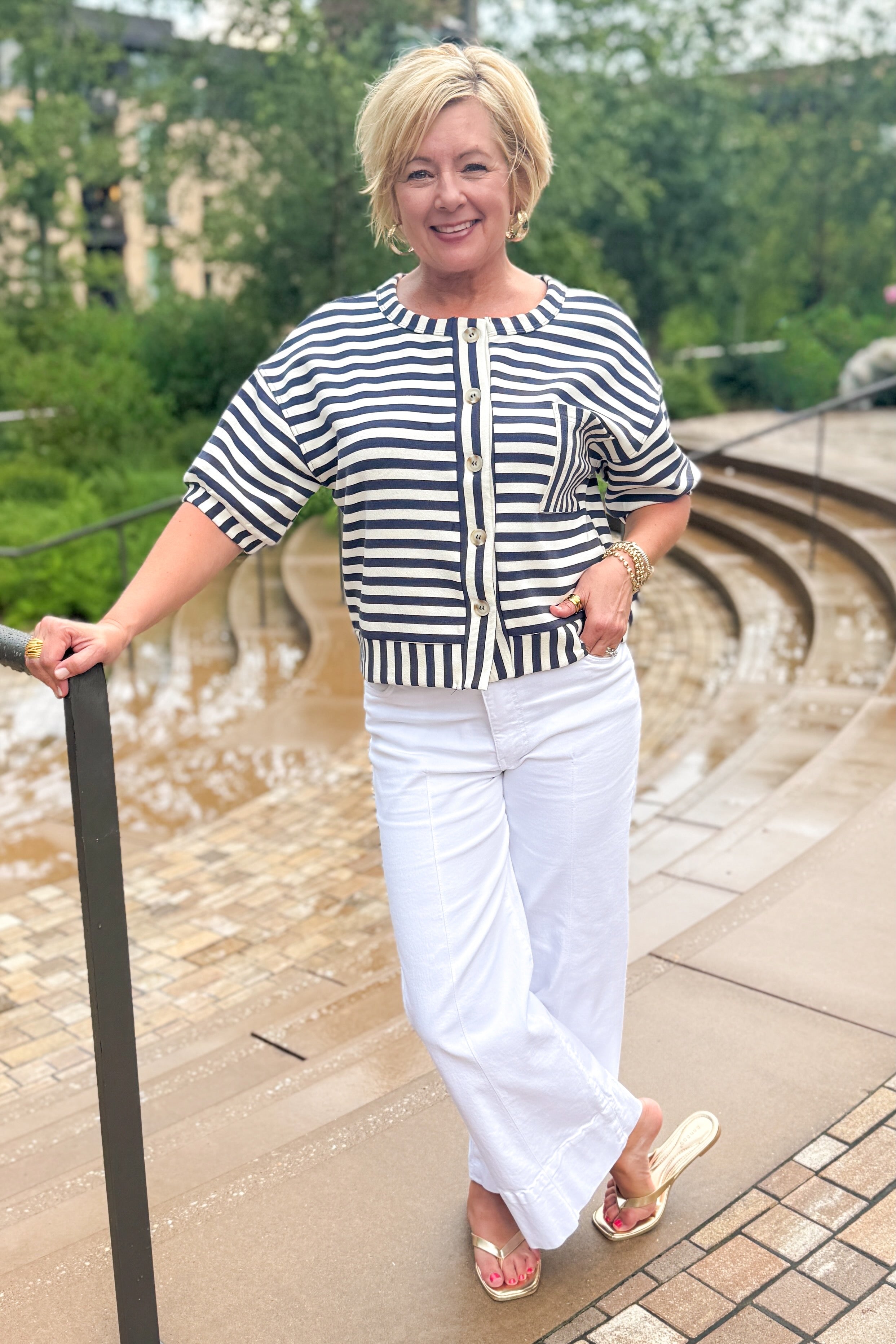 Front view of woman wearing Swagger Boutiqueβs Navy Striped Short Sleeve Button Up Top with jeans leaning against a railing outside.