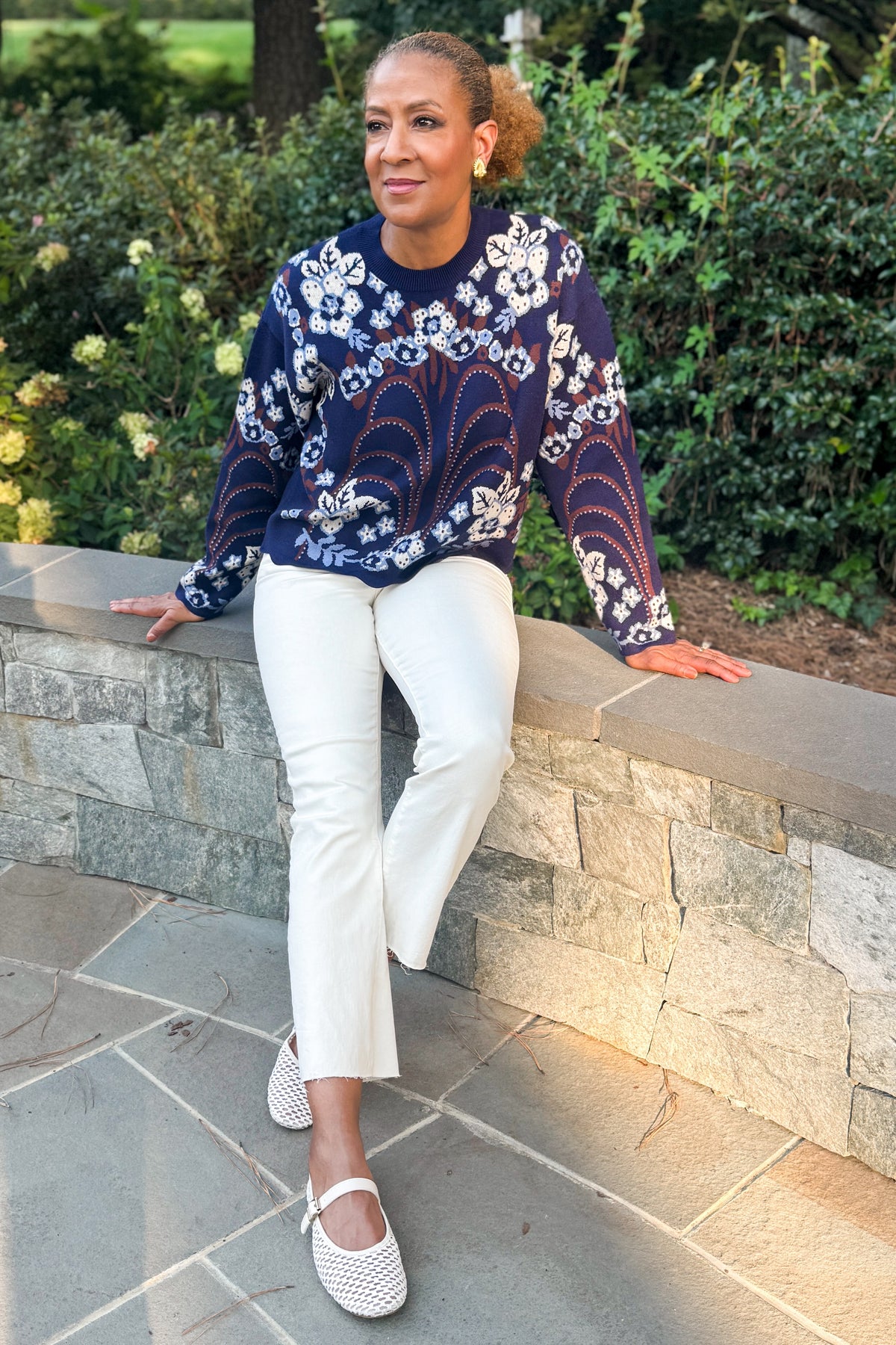 Front view of woman wearing Swagger Boutique’s Navy & Brown Printed Sweater by THML with jeans sitting on a stone wall outside.
