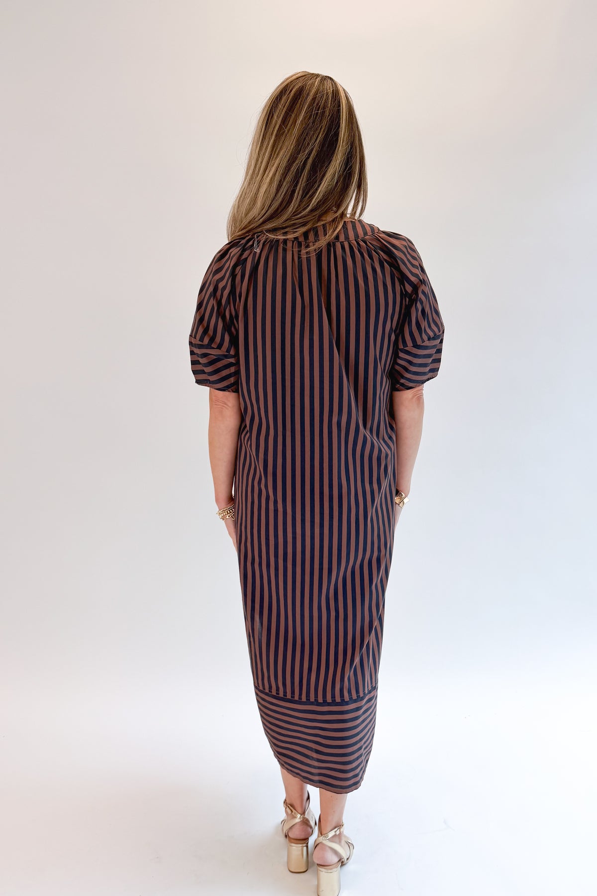 Back view of woman wearing Swagger Boutique’s Navy & Brown Striped Puff Sleeve Midi Dress by Entro with heels against white studio background.