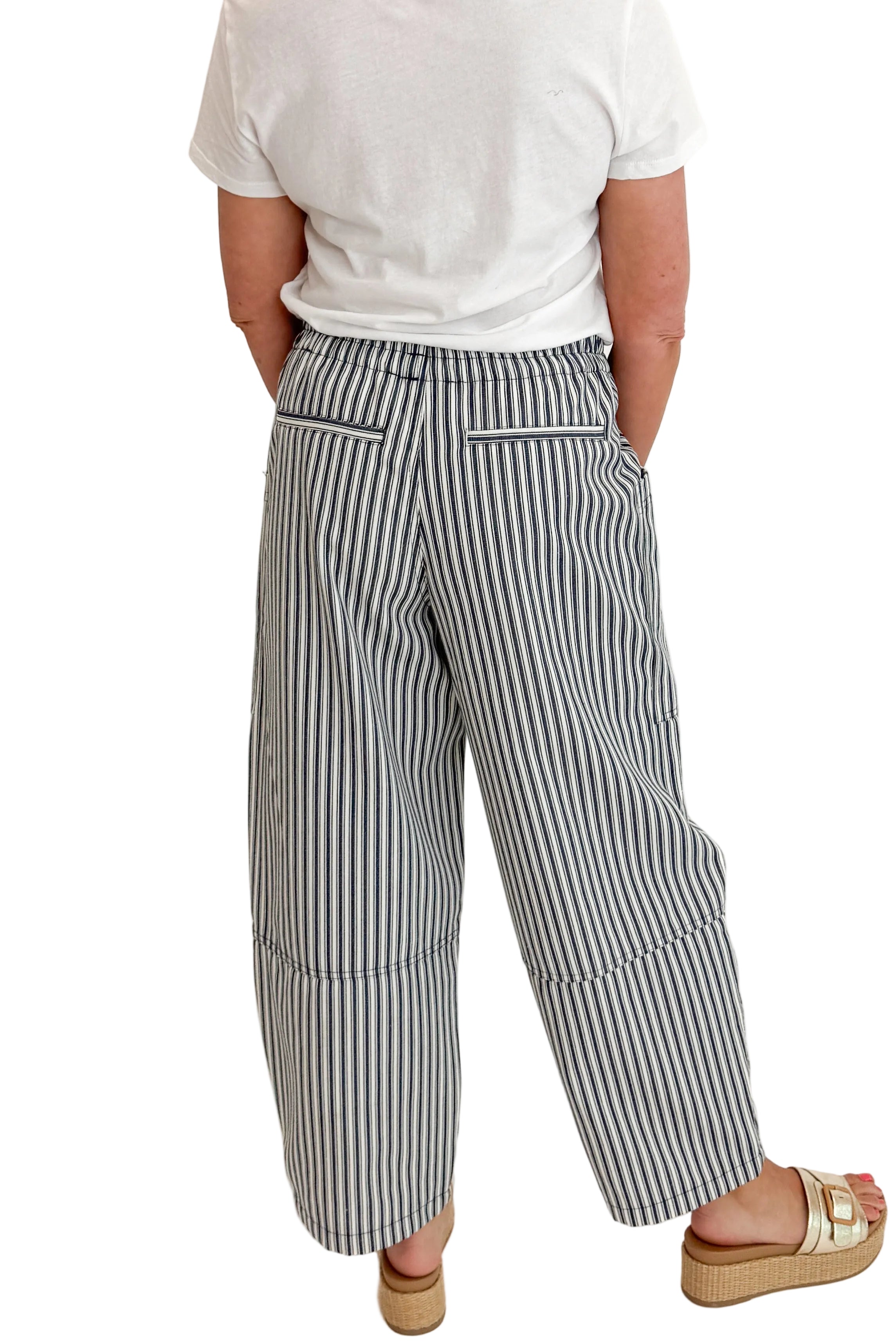 Back view of woman wearing Swagger Boutiqueβs In Full Swing Navy & Cream Striped Barrel Pants with white top and sandals against white studio background.