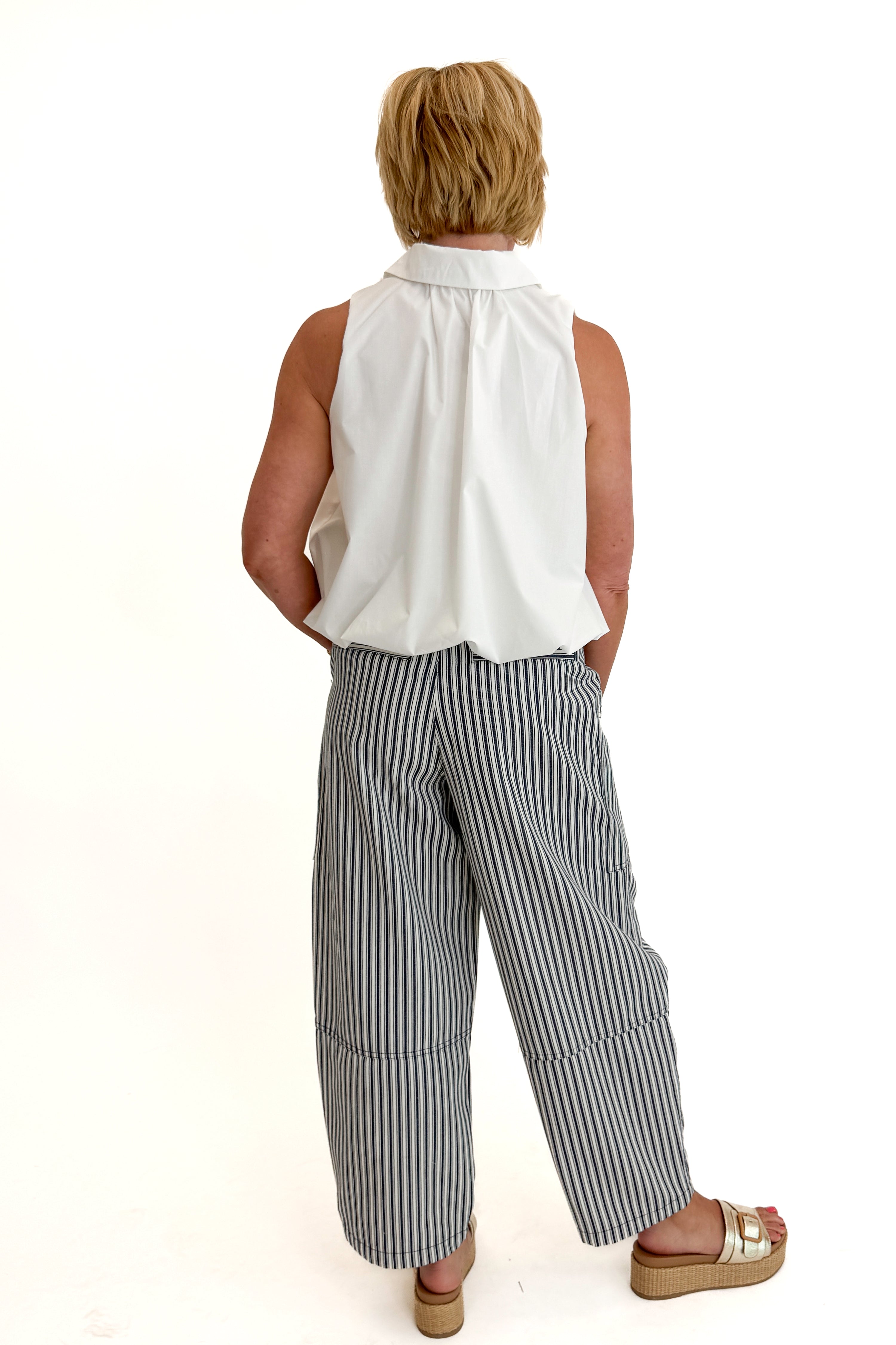 Back view of woman wearing Swagger Boutiqueβs In Full Swing Navy & Cream Striped Barrel Pants with white top and sandals against white studio background.
