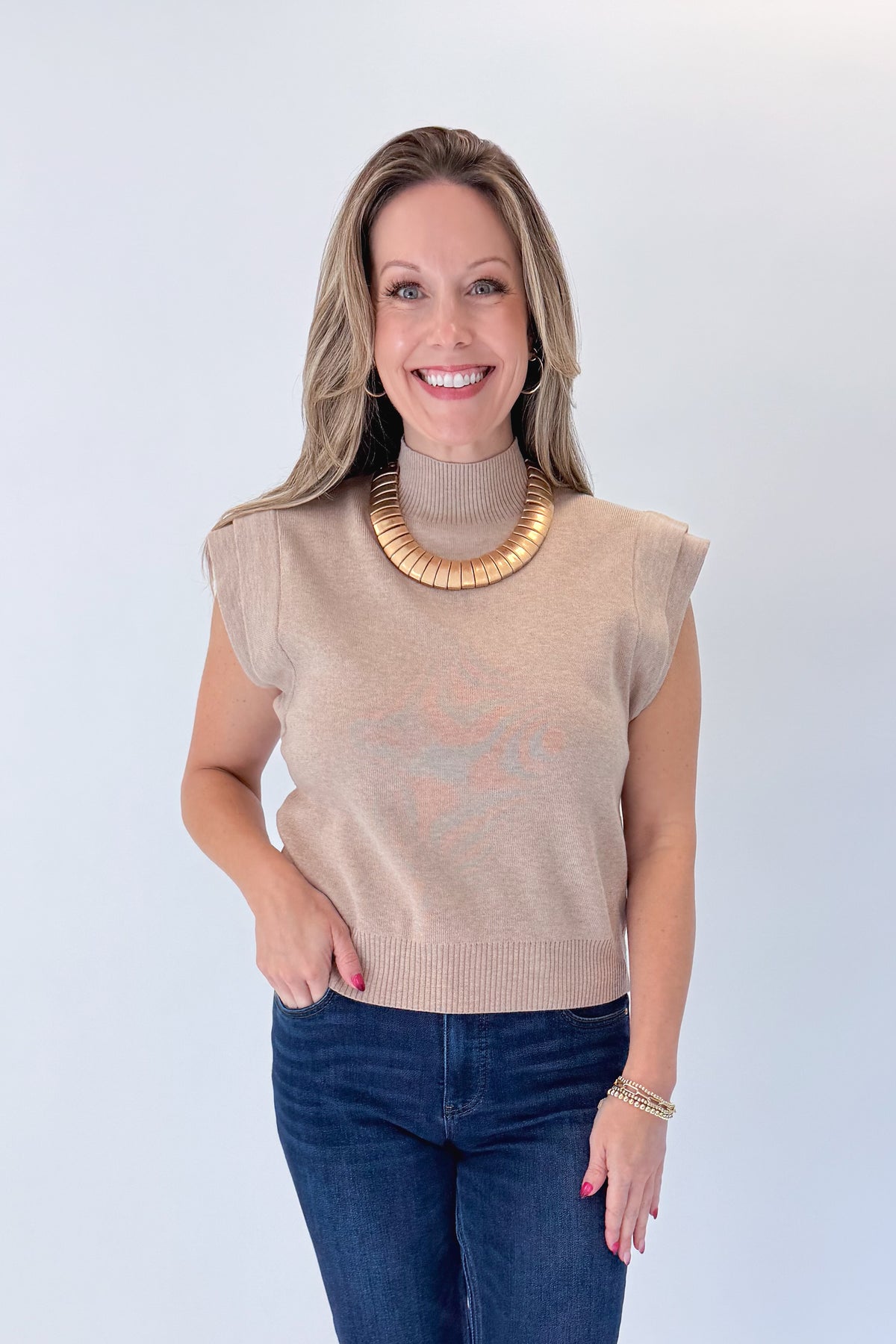 Front view of woman wearing Swagger Boutique’s Oatmeal Mock Neck Sleeveless Sweater by Entro with jeans against white studio background.