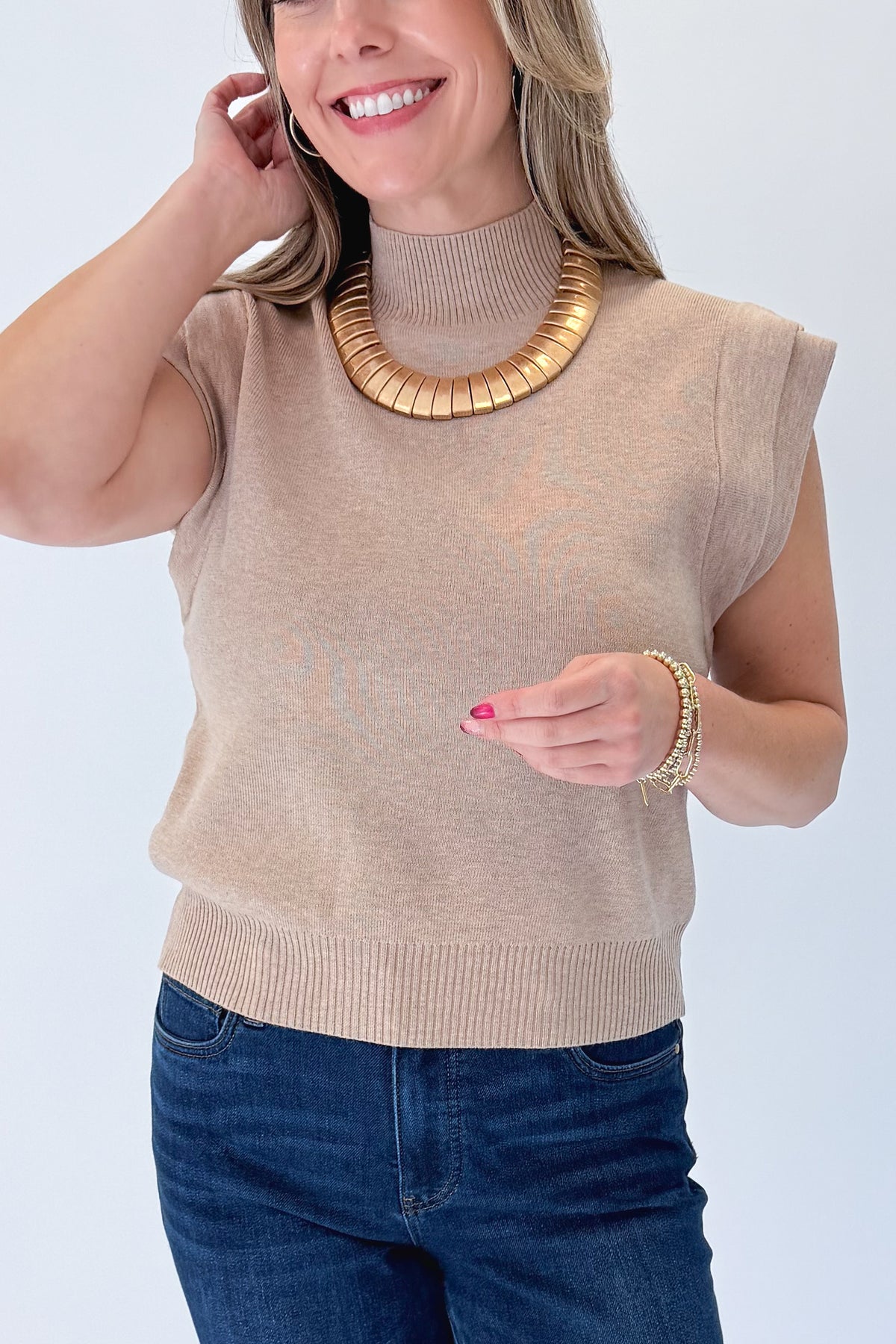 Front detail view of woman wearing Swagger Boutique’s Oatmeal Mock Neck Sleeveless Sweater by Entro with jeans against white studio background.