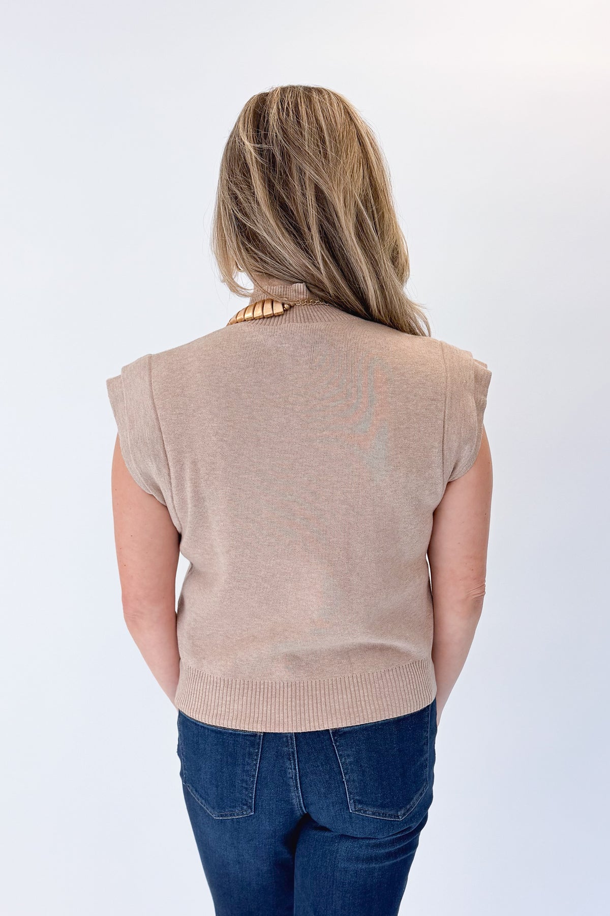 Back view of woman wearing Swagger Boutique’s Oatmeal Mock Neck Sleeveless Sweater by Entro with jeans against white studio background.