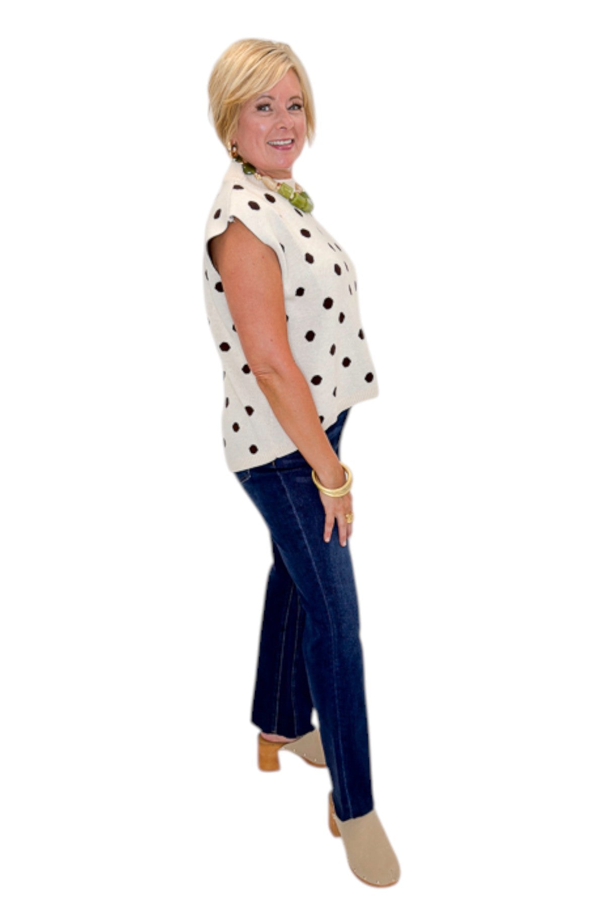 Side view of woman wearing Swagger Boutique’s Oatmeal & Brown Polka Dot Sleeveless Sweater by Entro with jeans against white studio background.