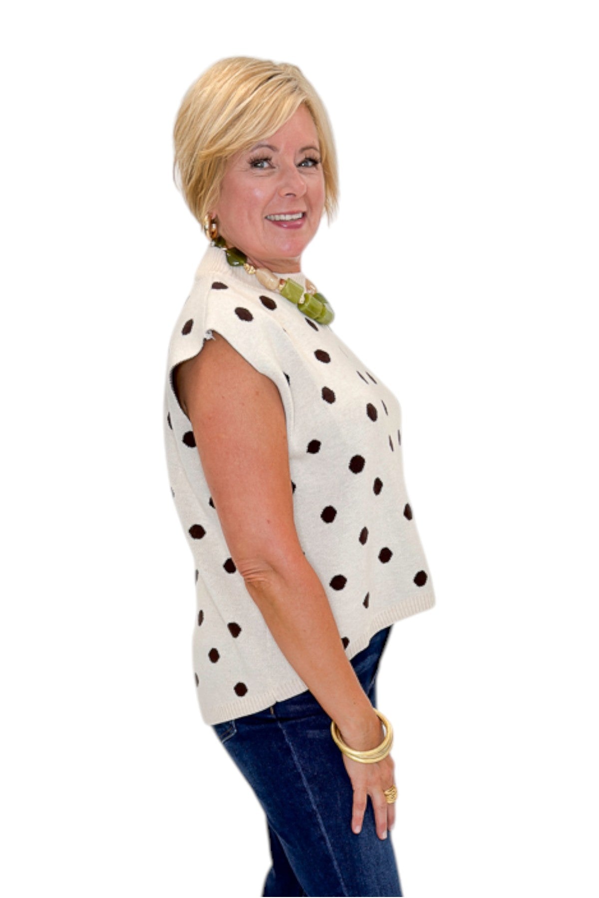 Side view of woman wearing Swagger Boutique’s Oatmeal & Brown Polka Dot Sleeveless Sweater by Entro with jeans against white studio background.