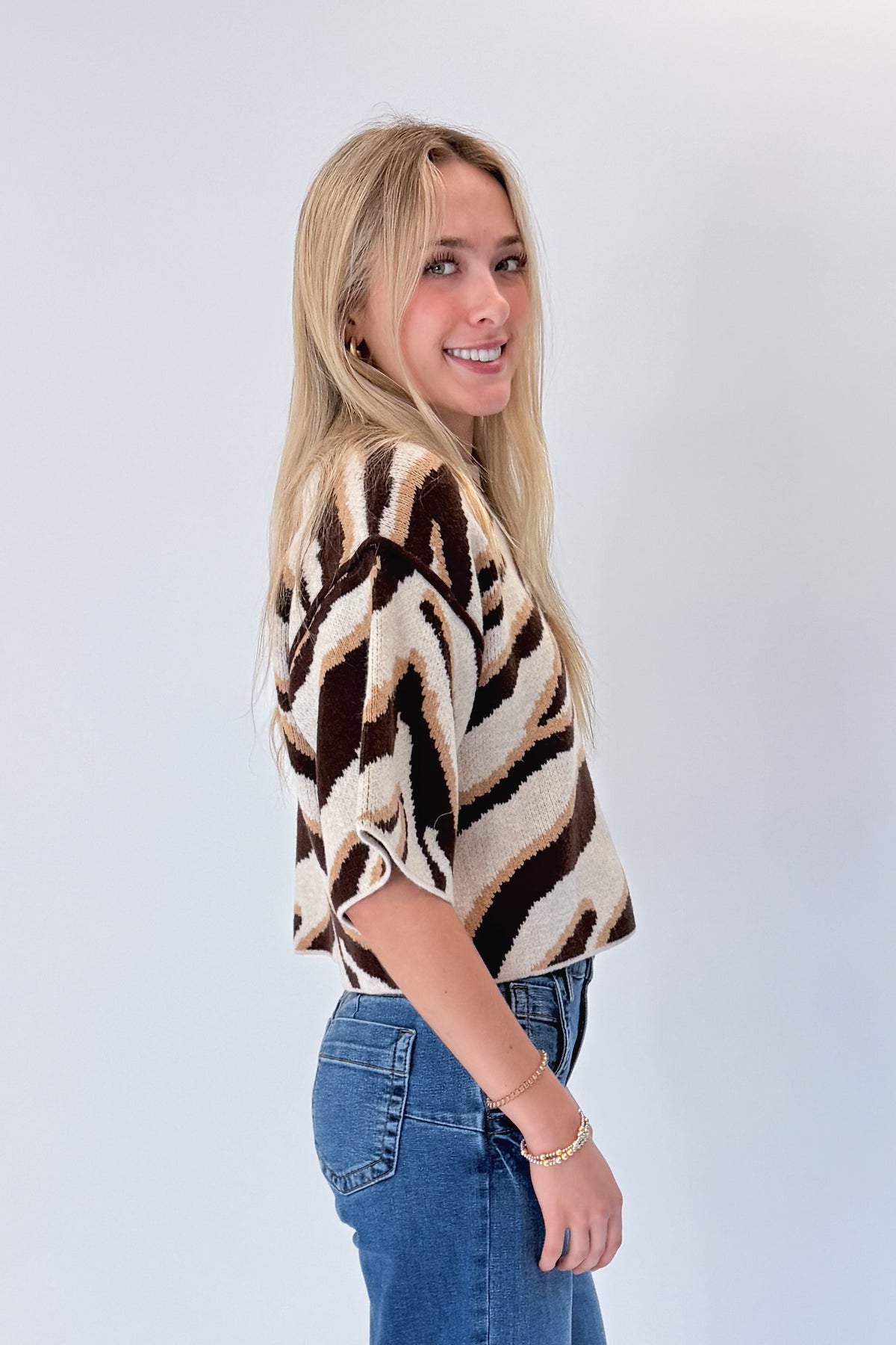 Side view of woman wearing Swagger Boutique’s Oatmeal & Brown Zebra Short Sleeve Sweater by Entro with jeans against white studio background.