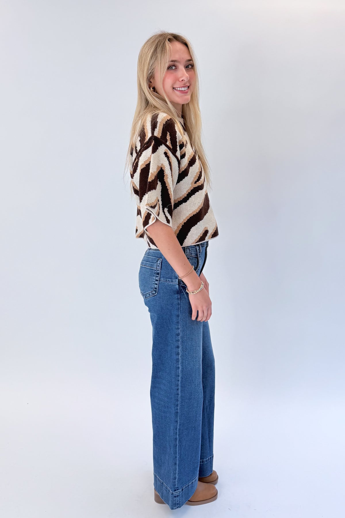 Side view of woman wearing Swagger Boutique’s Oatmeal & Brown Zebra Short Sleeve Sweater by Entro with jeans against white studio background.