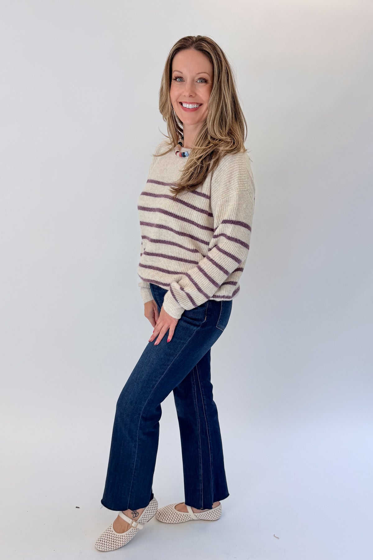 Side view of woman wearing Swagger Boutique’s Oatmeal & Purple Striped Sweater with jeans against white studio background.