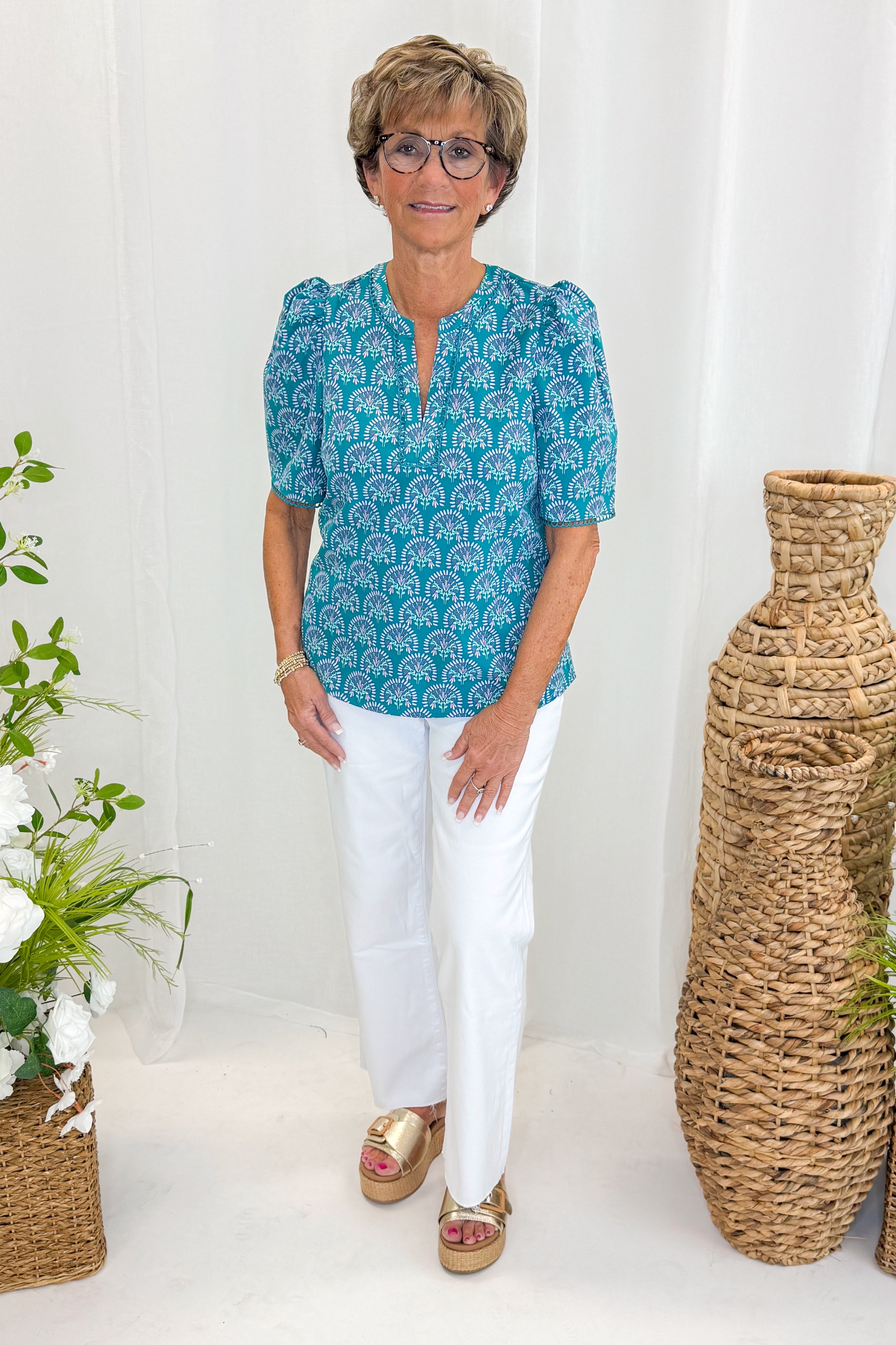 Front view of woman wearing Swagger Boutiqueβs Ocean Sunshower Flora Adelyn Top by Mary Square with jeans against decorative background.