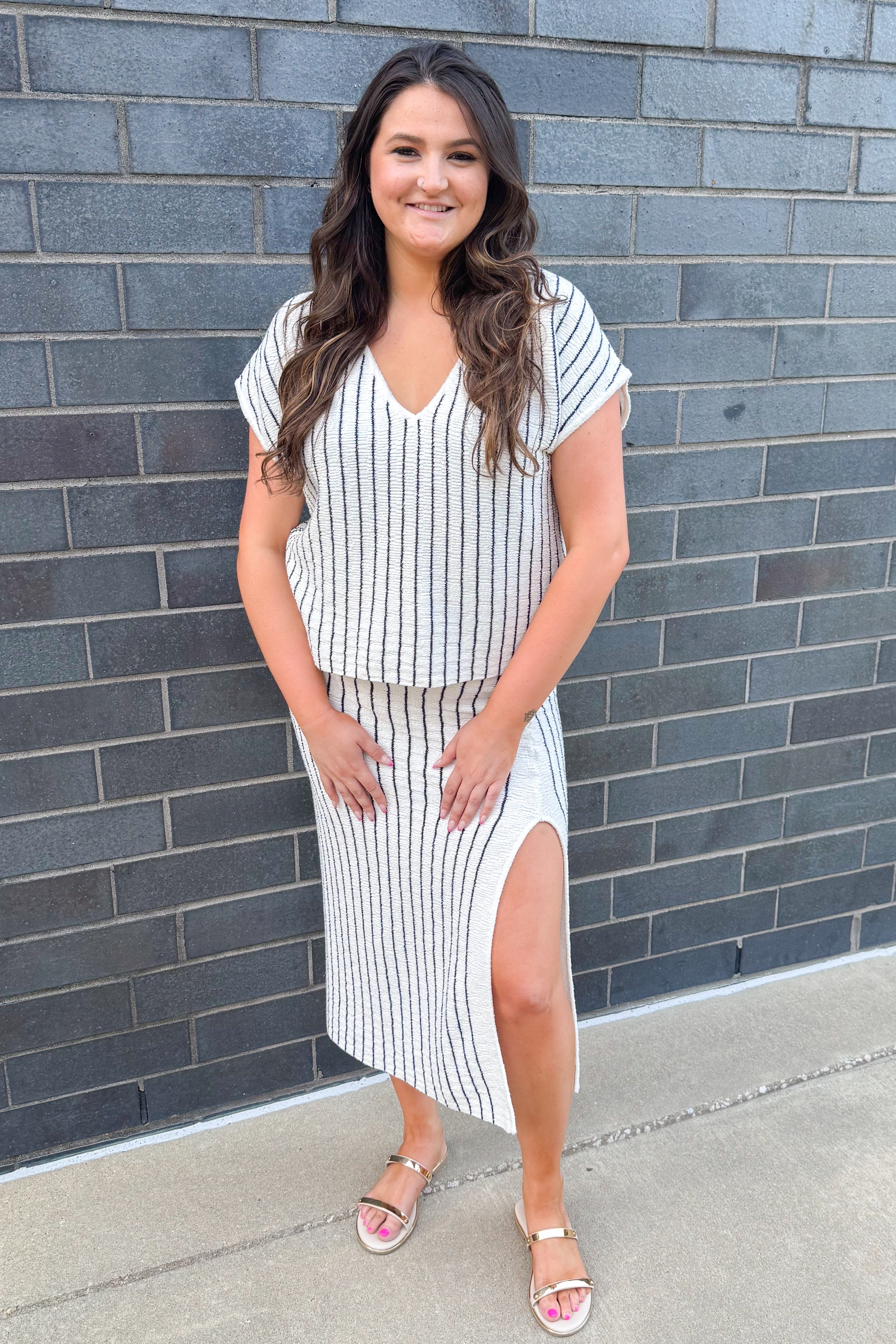 Front view of woman wearing Swagger Boutiqueβs Off White & Navy Striped V-neck Top with Off White & Navy Striped Midi Skirt With Slit outside against a brick wall.