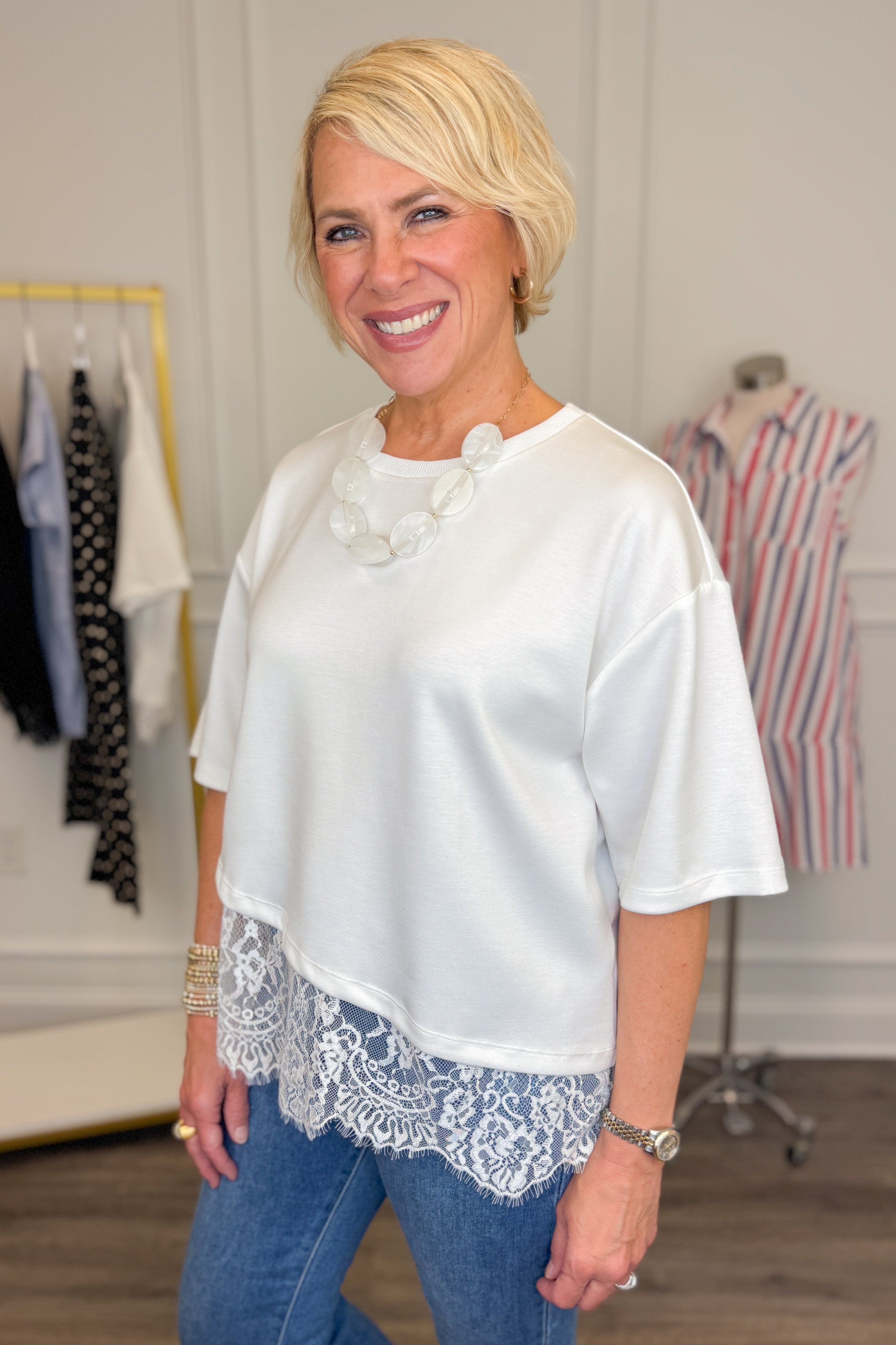Side view of woman wearing Swagger Boutiqueβs Off White Asymmetrical Lace Hem Top by Entro with jeans against white studio background.