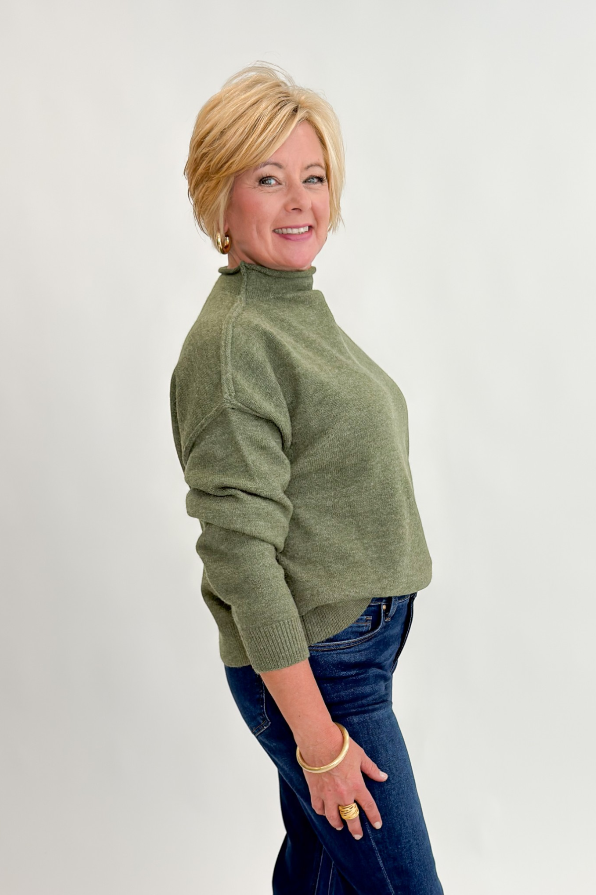 Side view of woman wearing Swagger Boutique’s Olive Mock Neck Sweater by Entro with jeans against white studio background.
