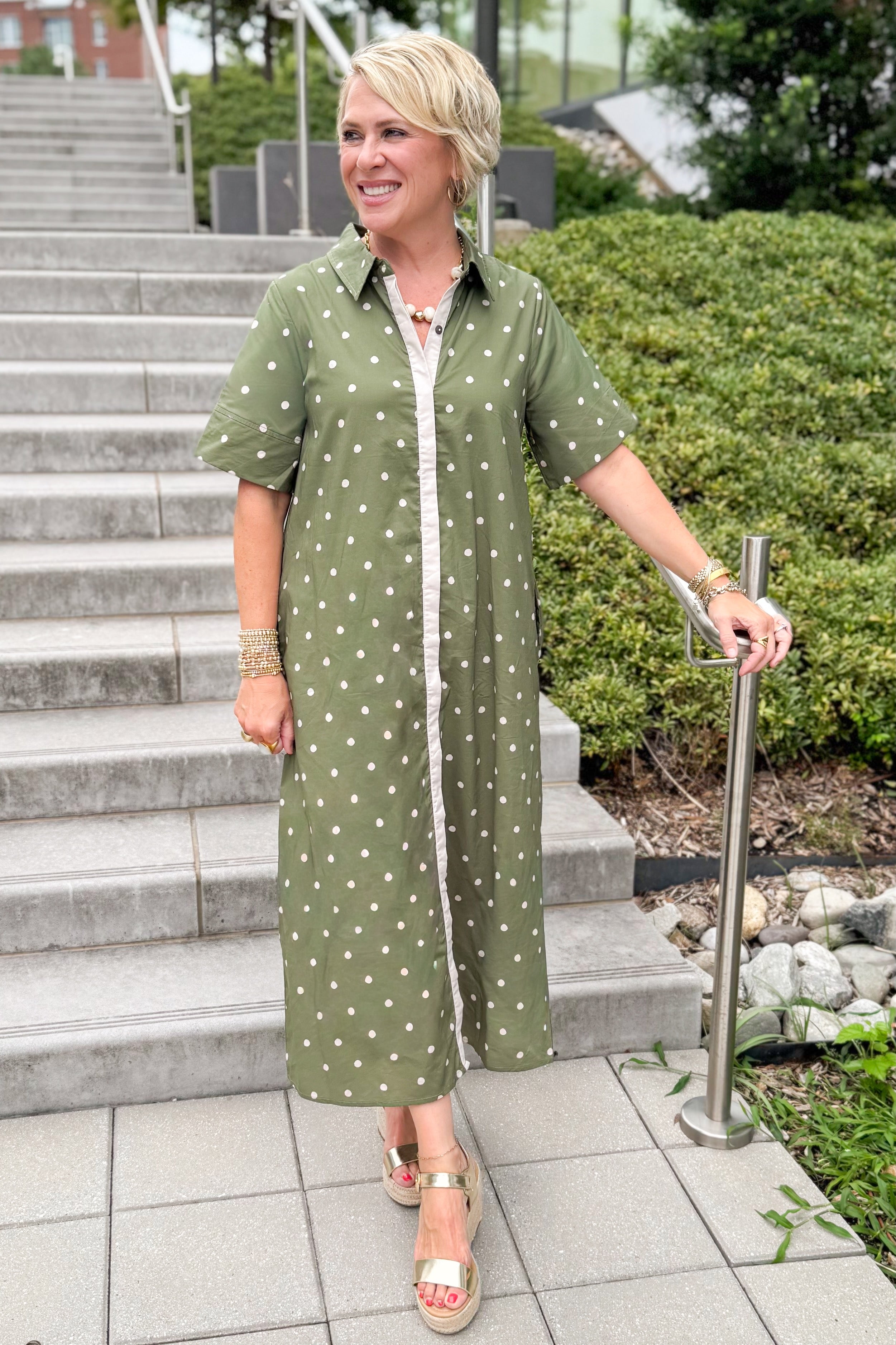Front view of woman wearing Swagger Boutiqueās Olive Polka Dot Midi Dress by Entro with sandals walking down a staircase outside.