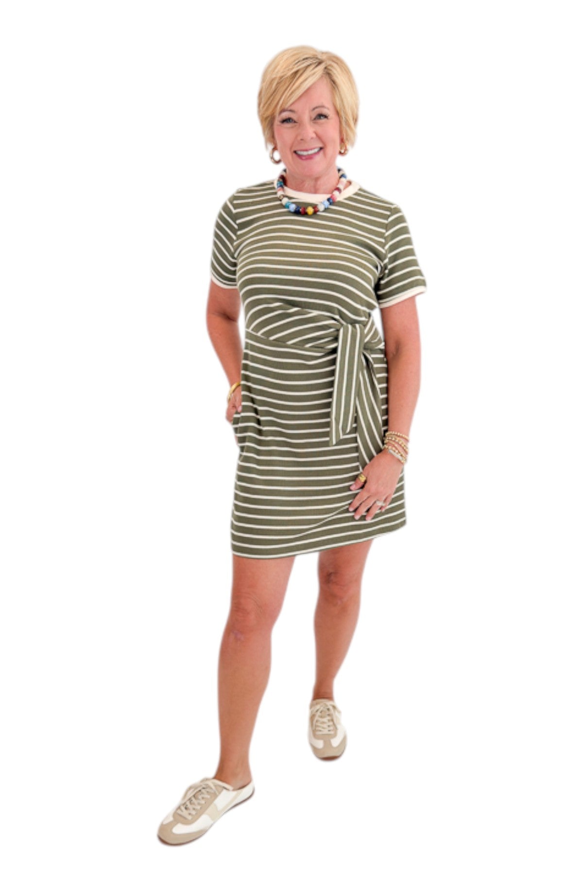 Front view of woman wearing Swagger Boutiqueās Olive Striped Tie Waist Mini Dress by Entro with sneakers against white studio background.