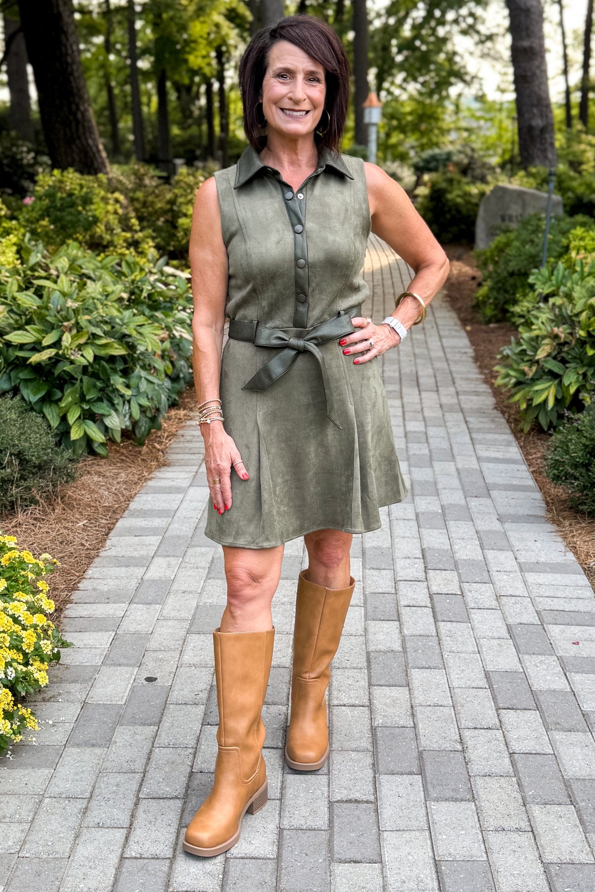 Front view of woman wearing Swagger Boutiqueโs Olive Suede Mini Dress with boots walking down a path outside.