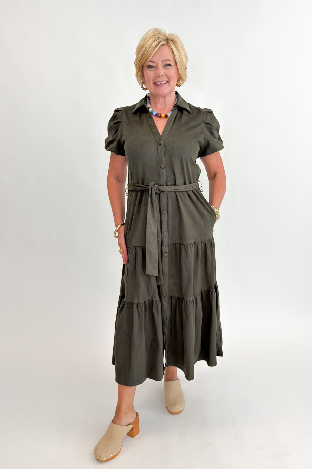 Front view of woman wearing Swagger Boutique’s Olive Suede Tiered Midi Dress with clogs against white studio background.