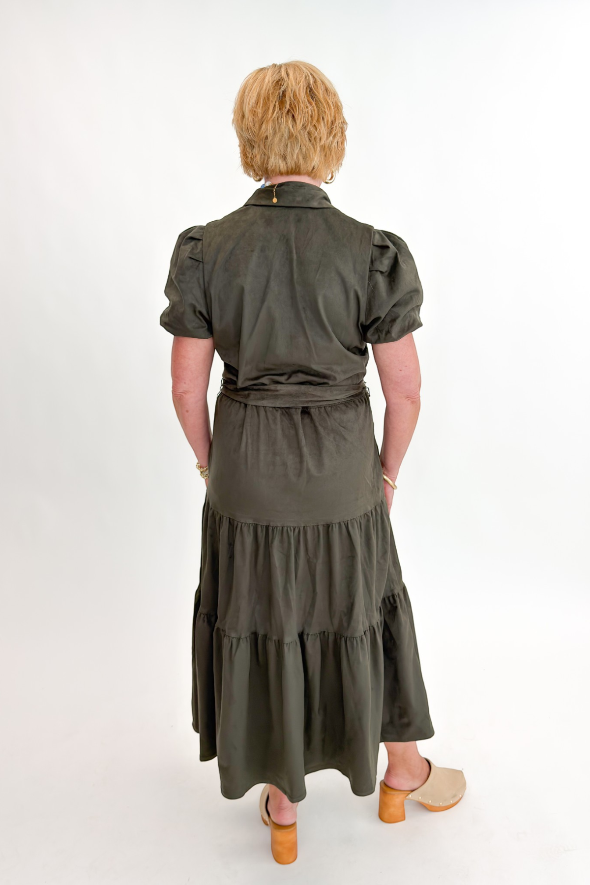 Back view of woman wearing Swagger Boutique’s Olive Suede Tiered Midi Dress with clogs against white studio background.