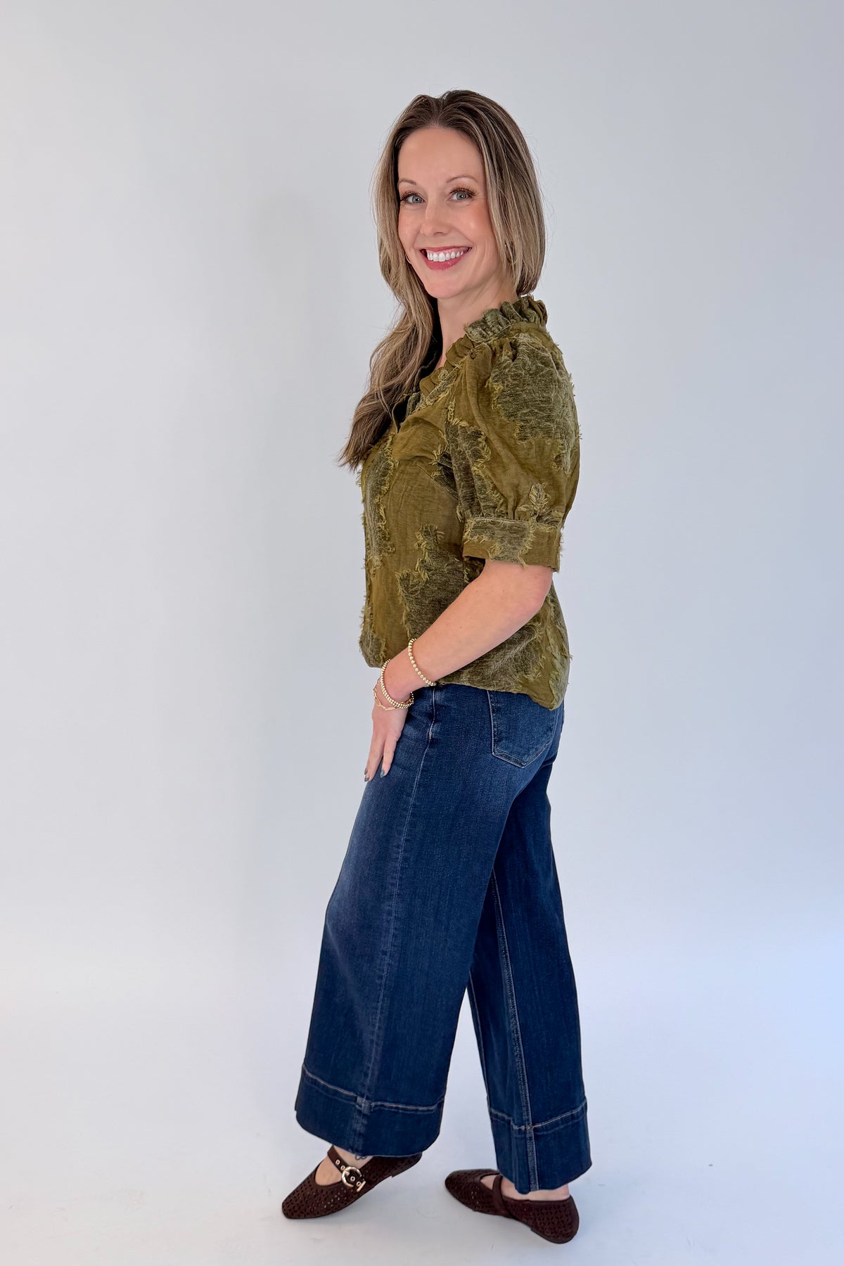 Side view of woman wearing Swagger Boutique’s Olive Velvet Ruffle Neck Top with jeans against white studio background.