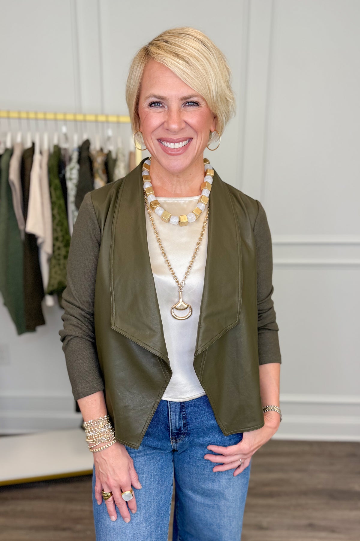 A smiling blonde woman stands indoors wearing an olive green wrap-style jacket over a white top and blue jeans, with layered gold and cream necklaces and bracelets. A clothing rack is visible in the softly lit boutique-like background.