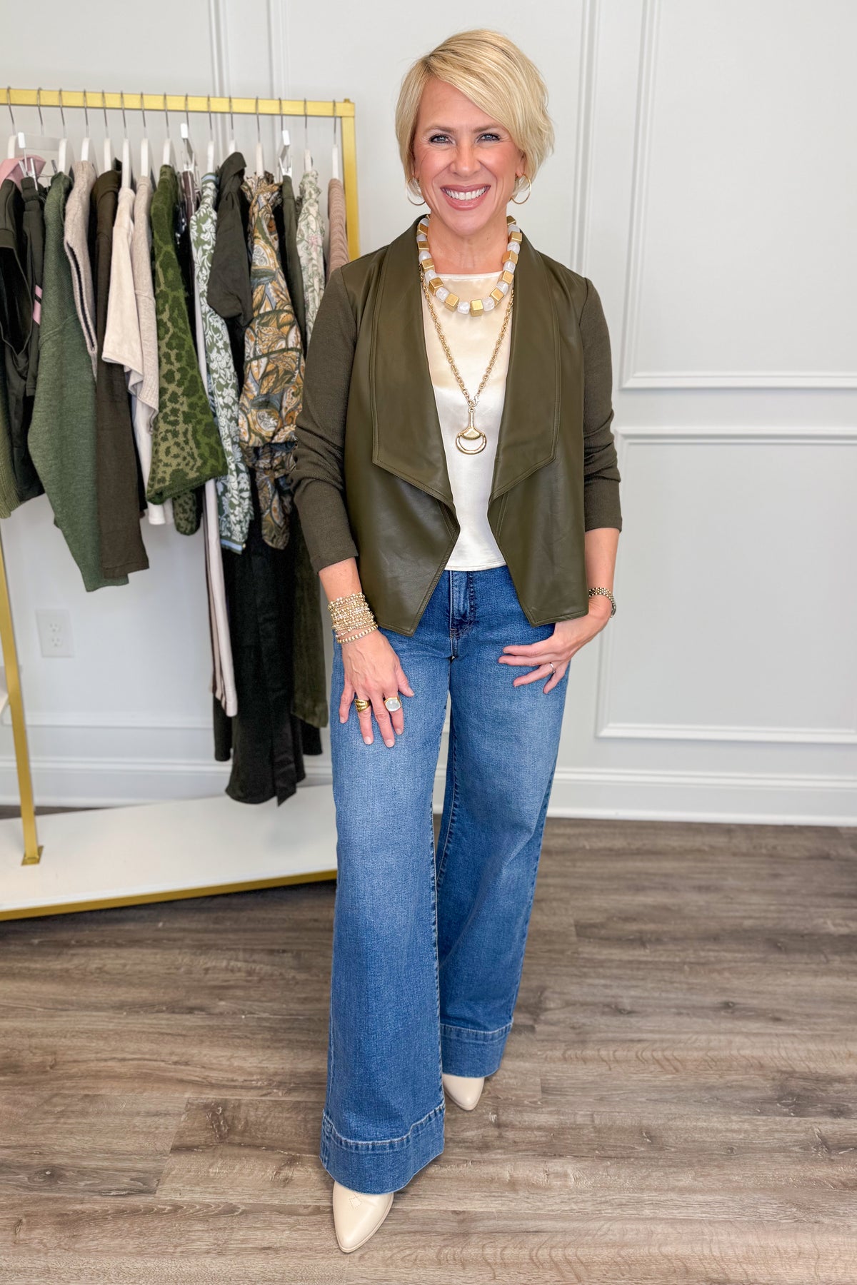 A woman stands in a boutique wearing an olive green wrap-style jacket, a white top, wide-leg blue jeans, and cream pointed-toe heels. A clothing rack with assorted garments is visible behind her against a white paneled wall.