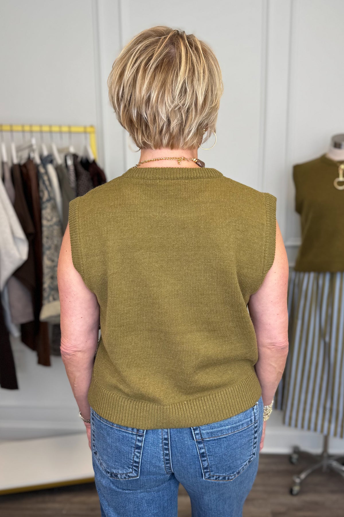 Back view of woman wearing Swagger Boutique’s Olive Sleeveless Sweater with jeans with clothing hanging in the background.