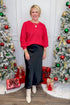 Front view of woman wearing red boatneck sweater with black skirt with Christmas trees in the background.