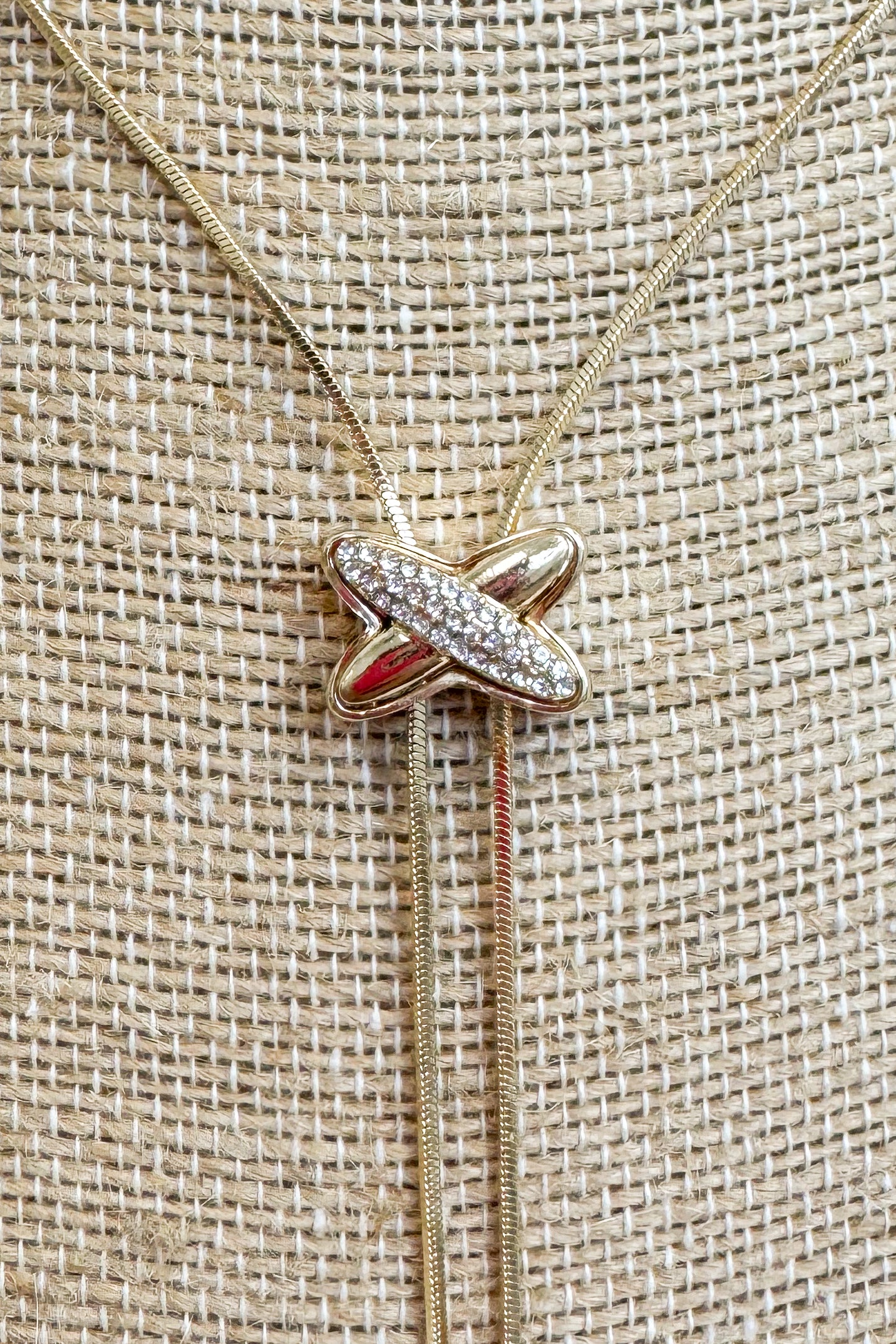 Close up of Gold-tone delicate chain necklace featuring a elongated vertical X pendant on a beige jewelry bust