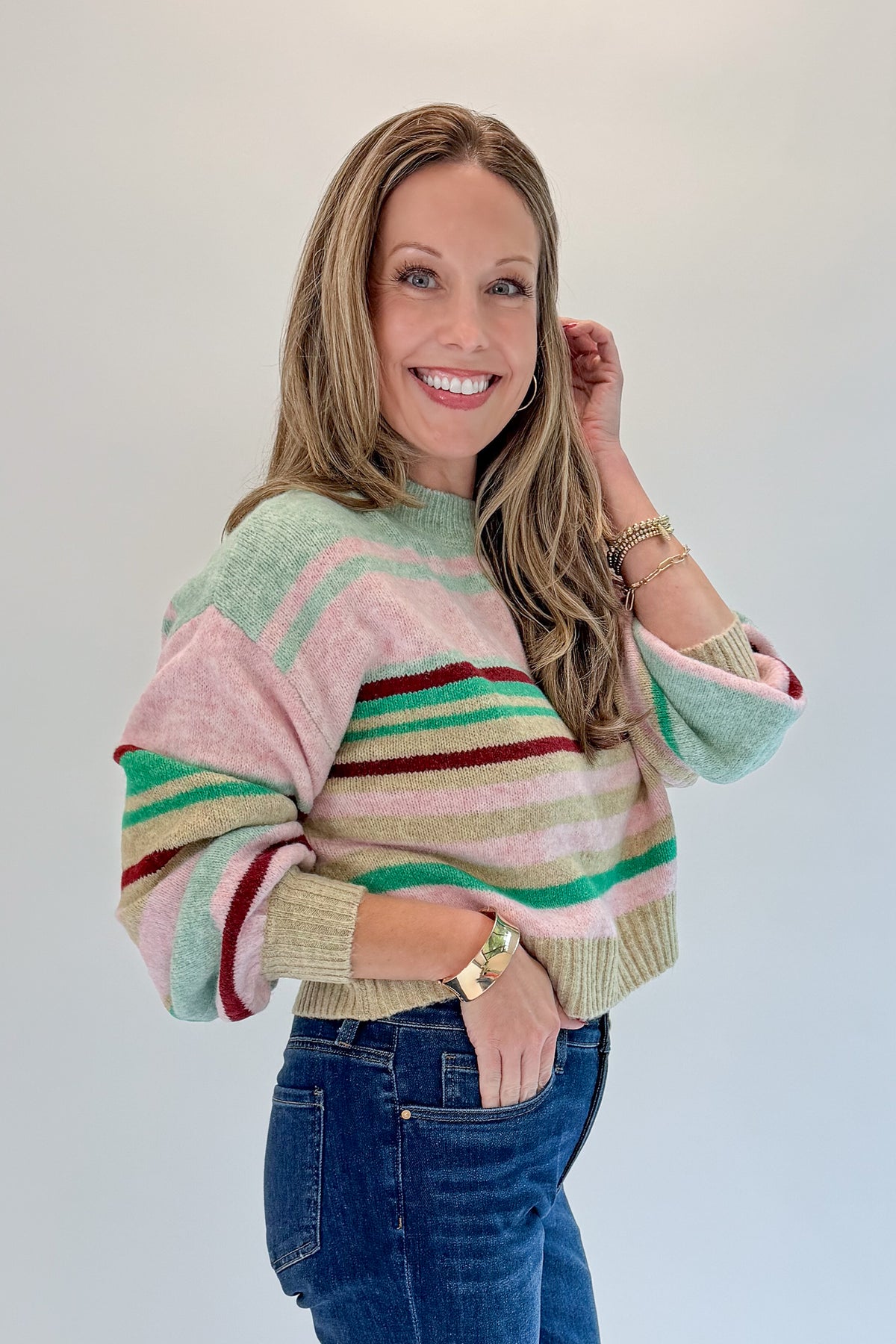 Side view of woman wearing Swagger Boutique’s Pink & Multi Striped Sweater with jeans against white studio background.