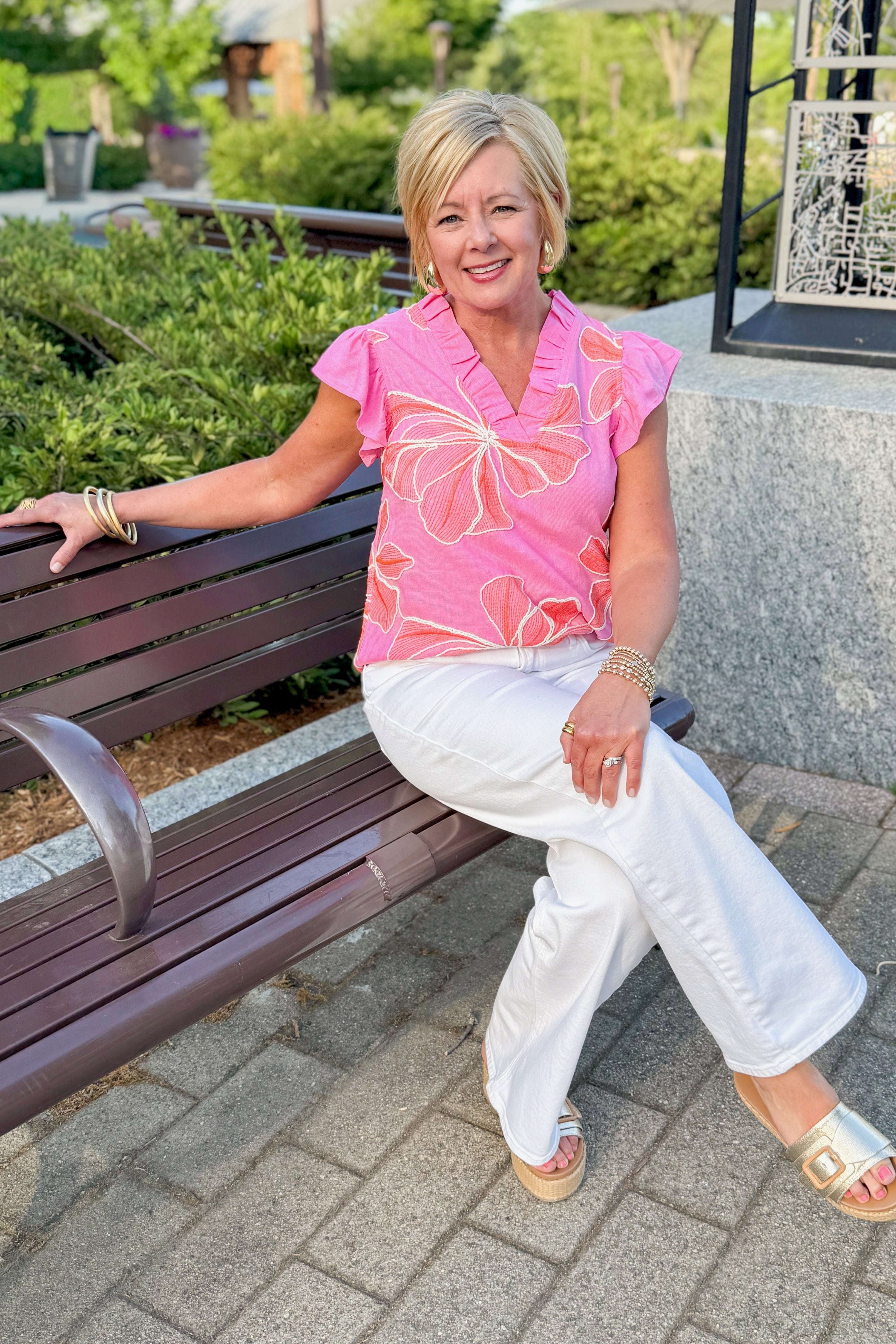 Front view of woman wearing Pink & Orange Floral Print Top with jeans sitting outside on a bench.