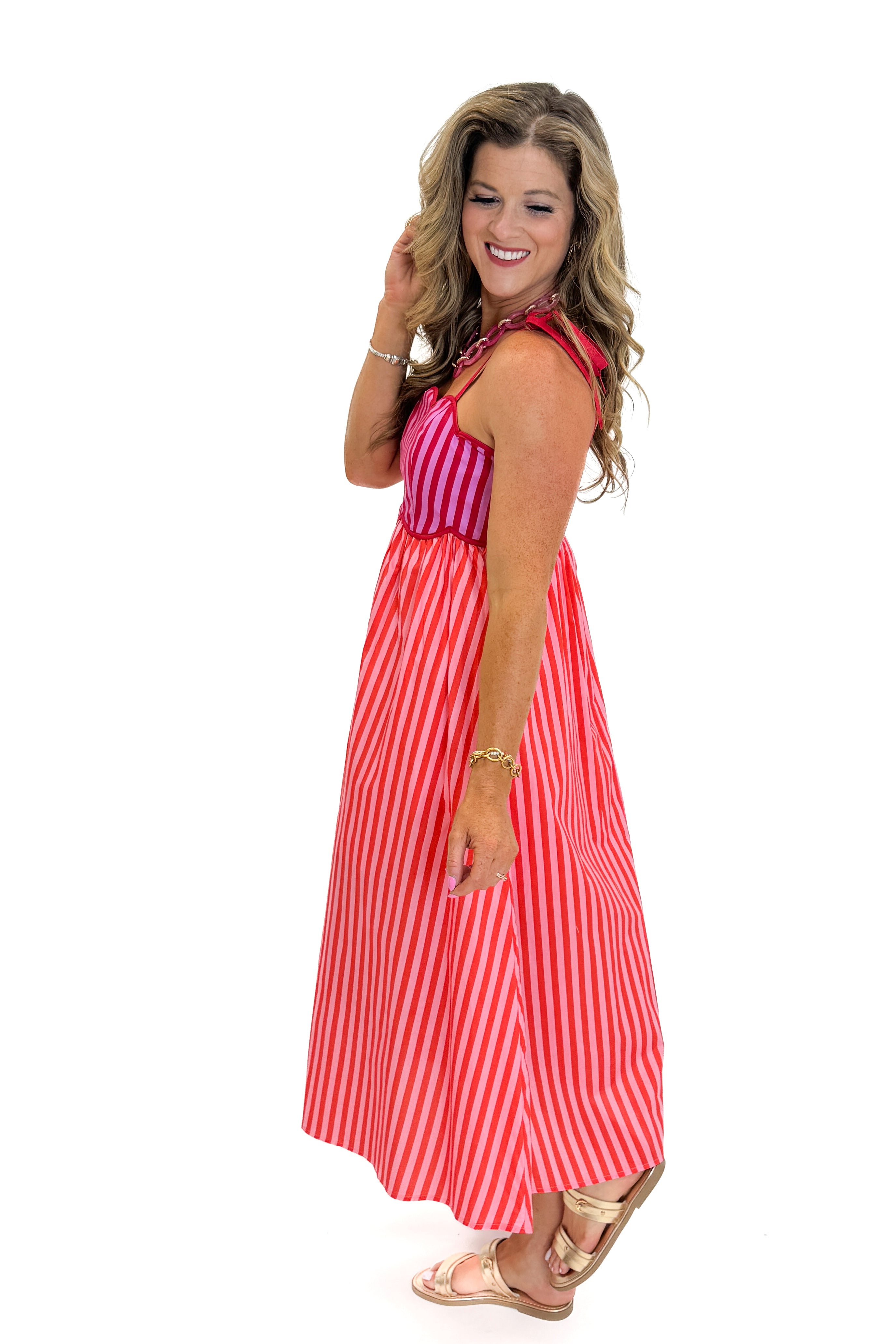 Side view of woman wearing Swagger Boutiqueβs Pink & Red Striped Scalloped Midi Dress by Entro with sandals against white studio background.