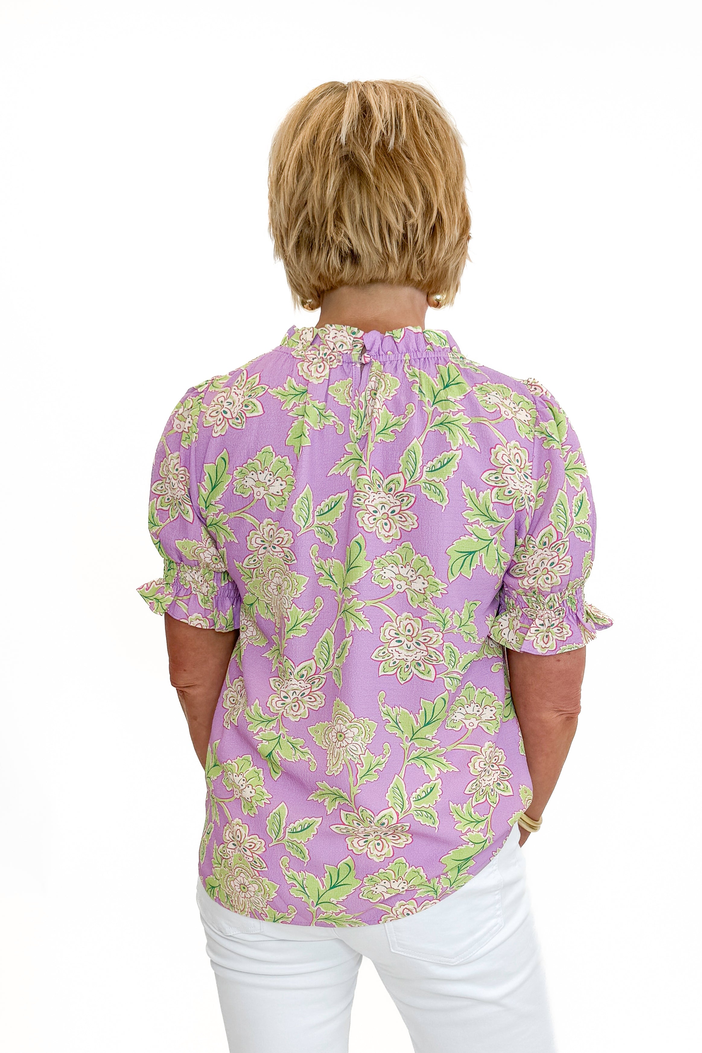 Back view of woman wearing Swagger Boutiqueโs Purple Floral Short Sleeve Ruffle Top with jeans against white studio background.
