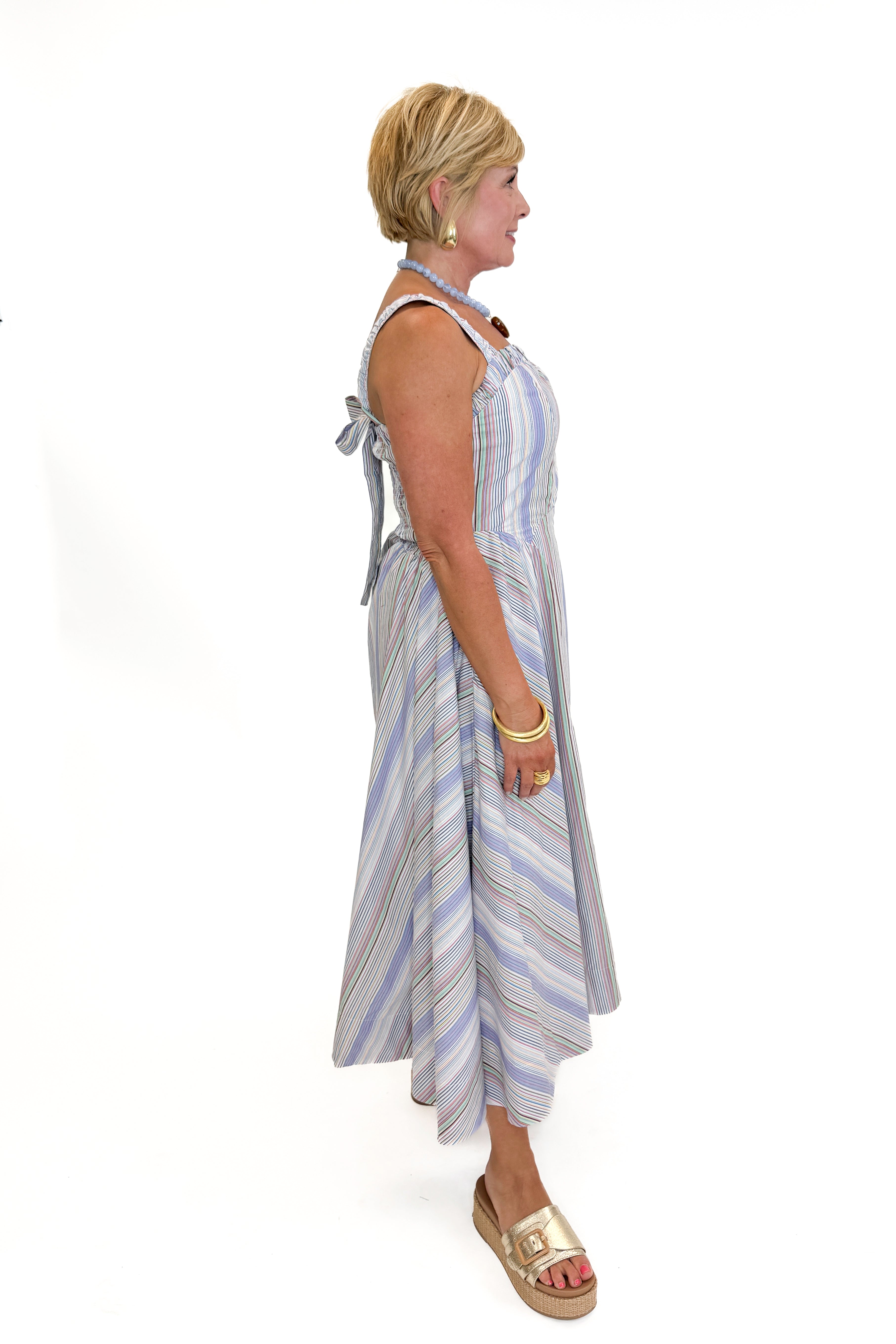 Side view of woman wearing Swagger Boutique’s Purple & Multi Striped Sleeveless Midi Dress with sandals against white studio background.