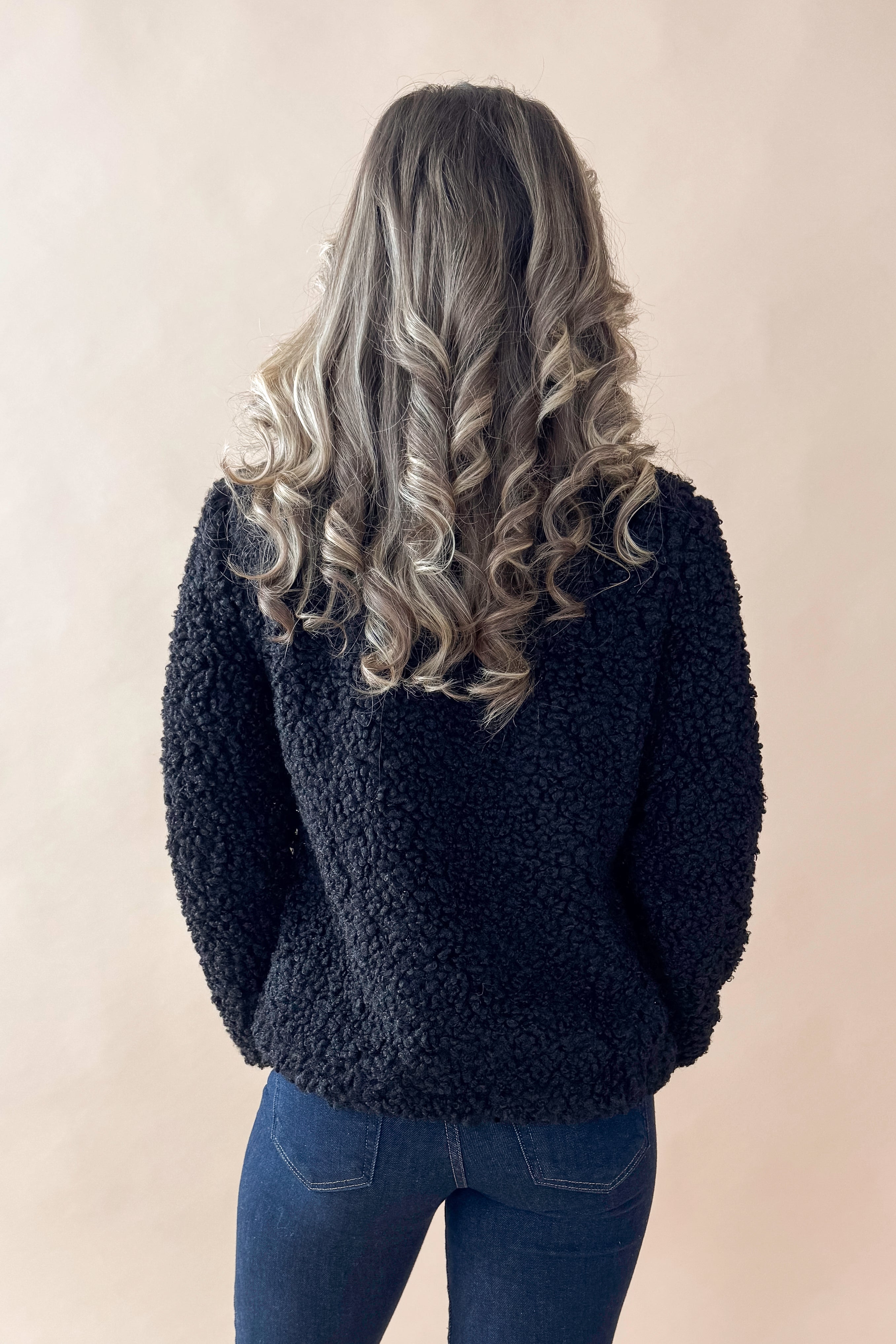 Back view of woman wearing Black Sherpa Coat and jeans against tan studio background.