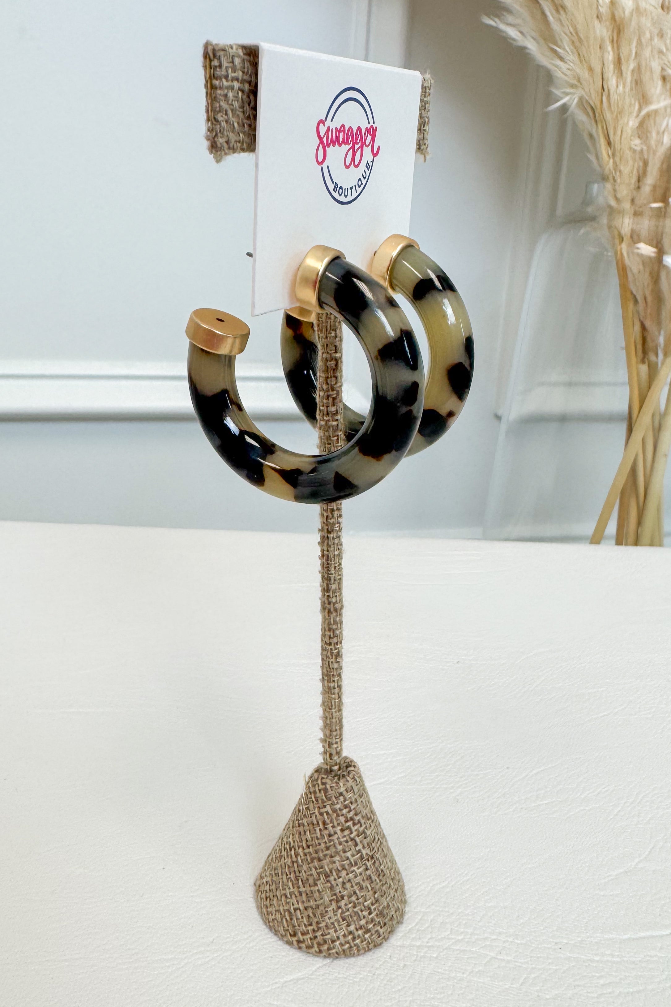 A pair of large tortoiseshell hoop earrings with gold-tone tips, displayed on a burlap-covered stand with a white card