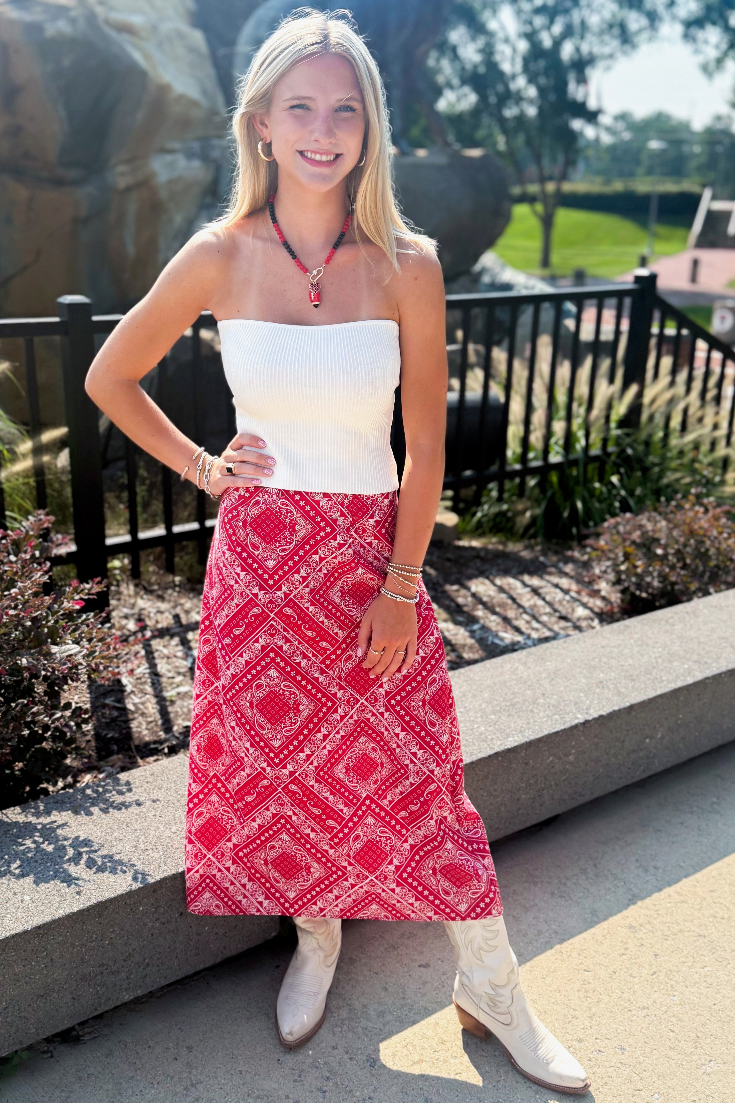 Front view of woman wearing Swagger Boutiqueās Red Bandana Print Midi Skirt with white top standing outside at a park.