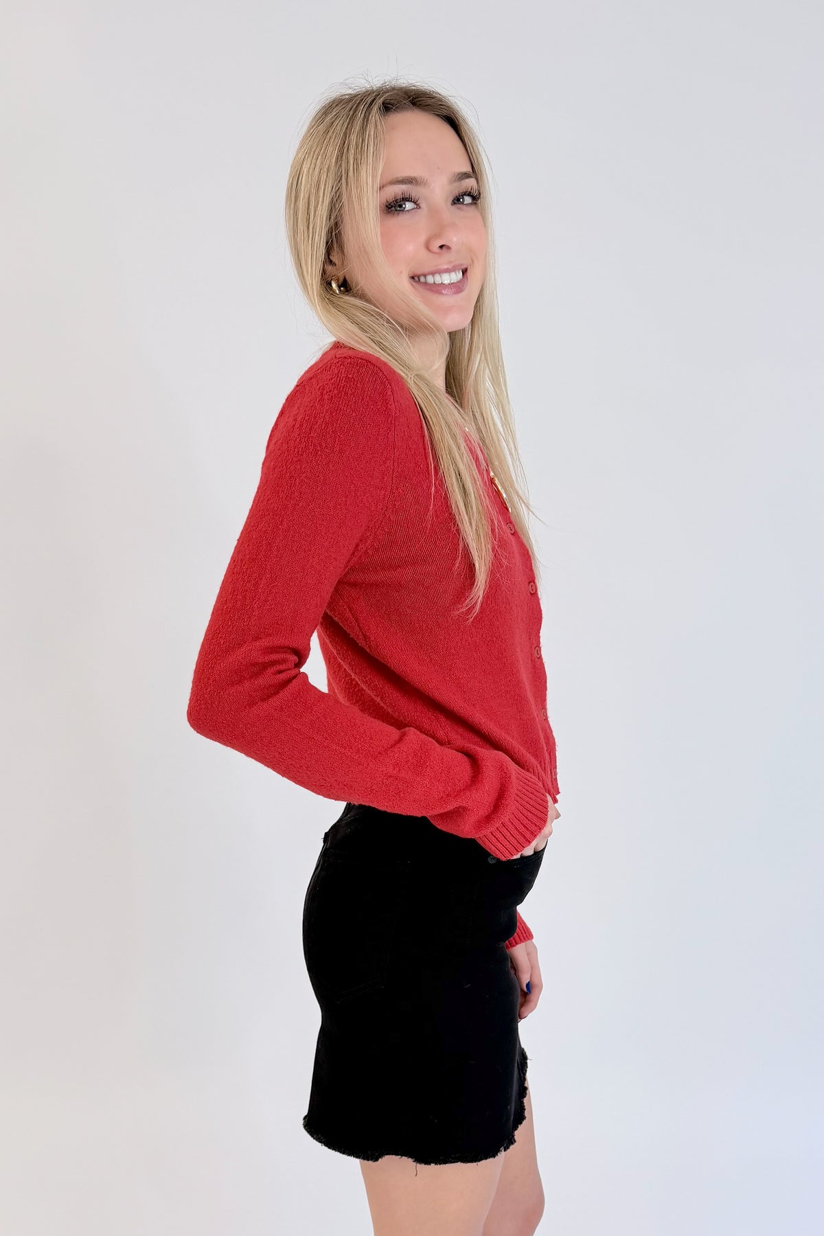 Side view of woman wearing Swagger Boutique’s Red Button Up Cardigan with Black Denim Mini Skirt by Just Black Denim against white studio background.