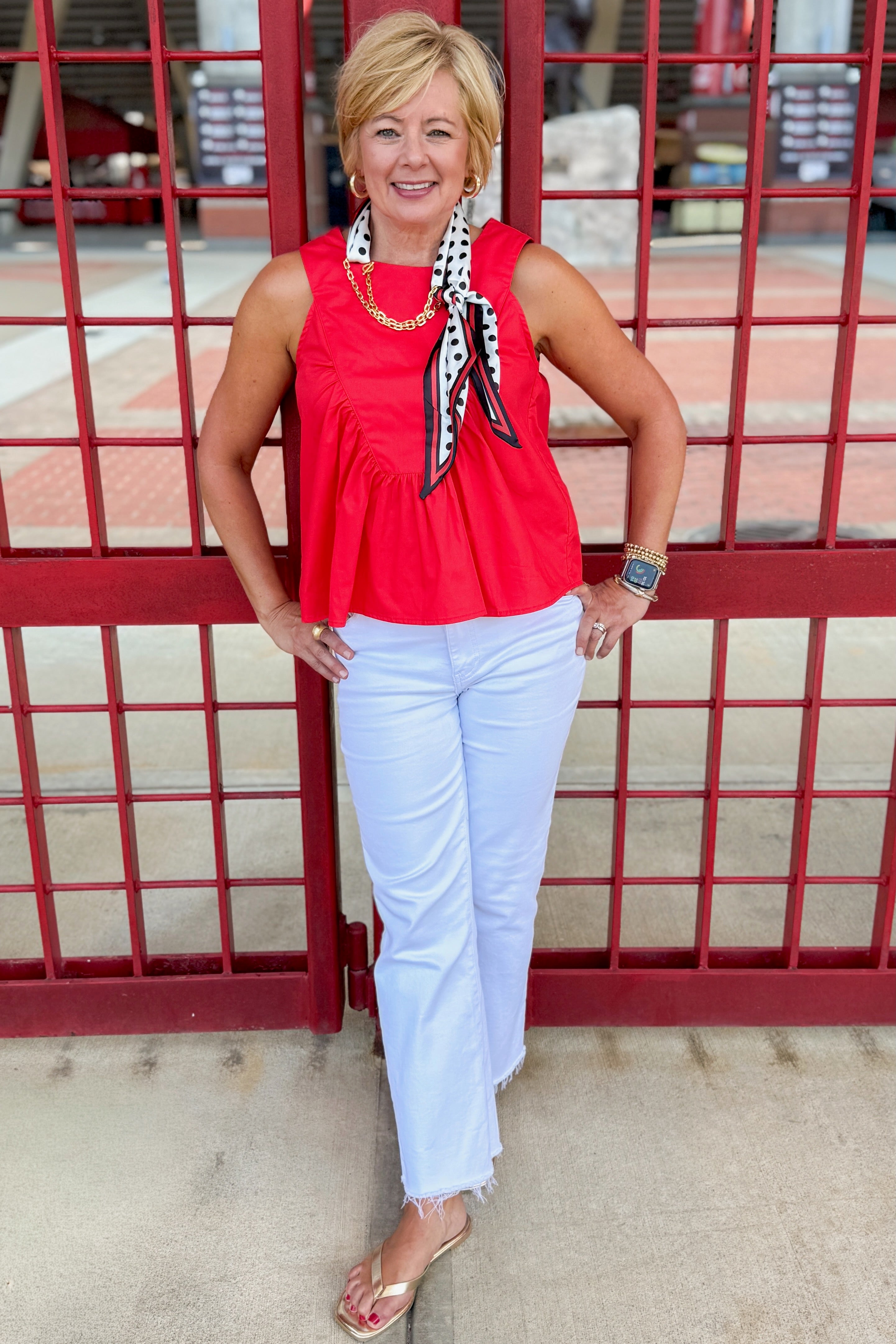 Front view of woman wearing Swagger Boutique’s Red Ruffle Tank with jeans standing by a stadium gate.