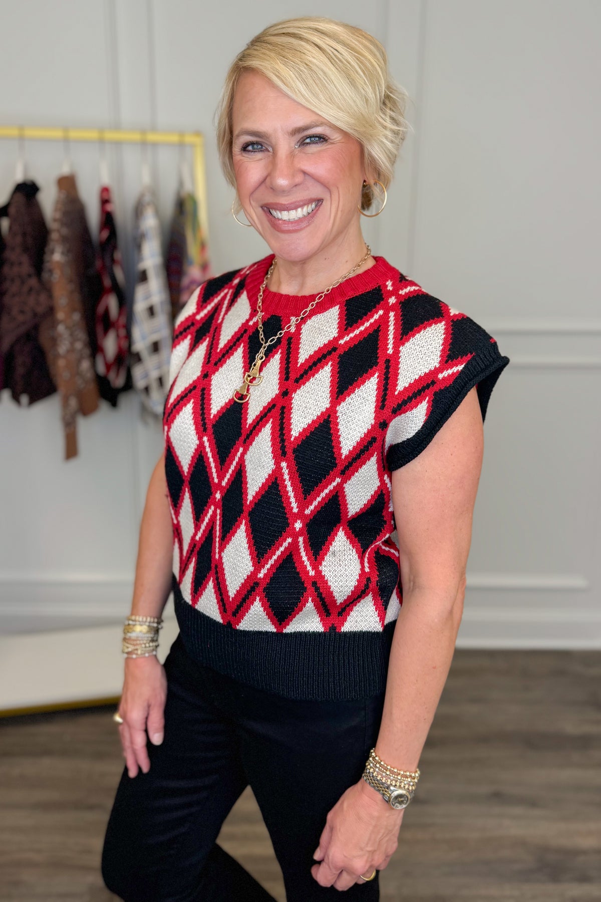 A smiling blonde woman wearing a red, black, and white argyle knit sweater vest with black pants, standing in a boutique setting with clothing racks visible in the background.