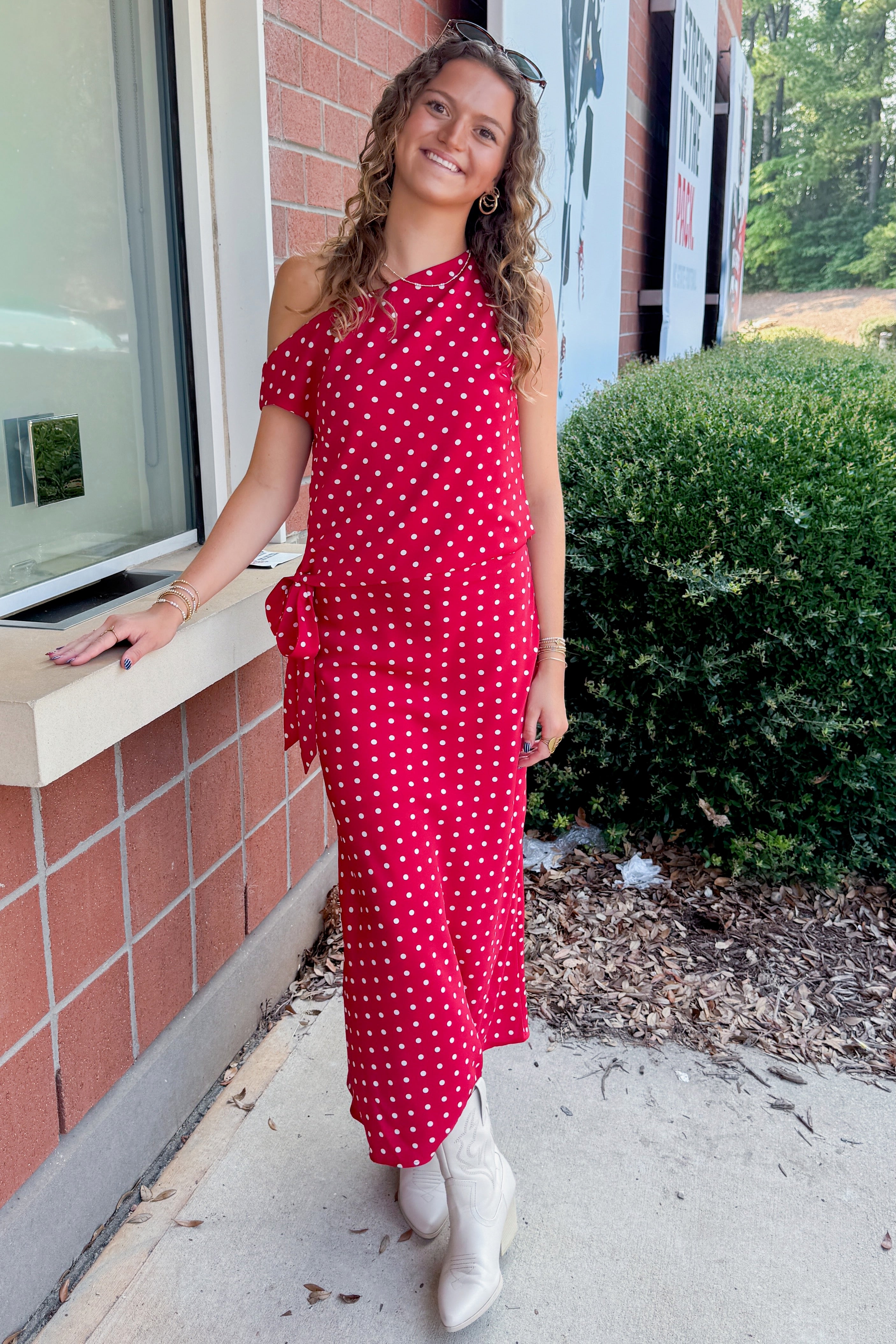 Front view of woman wearing Swagger Boutique’s Red & White Polka Dot Midi Skirt with Red & White Polka Dot Asymmetrical Tank standing outside a ticket office.