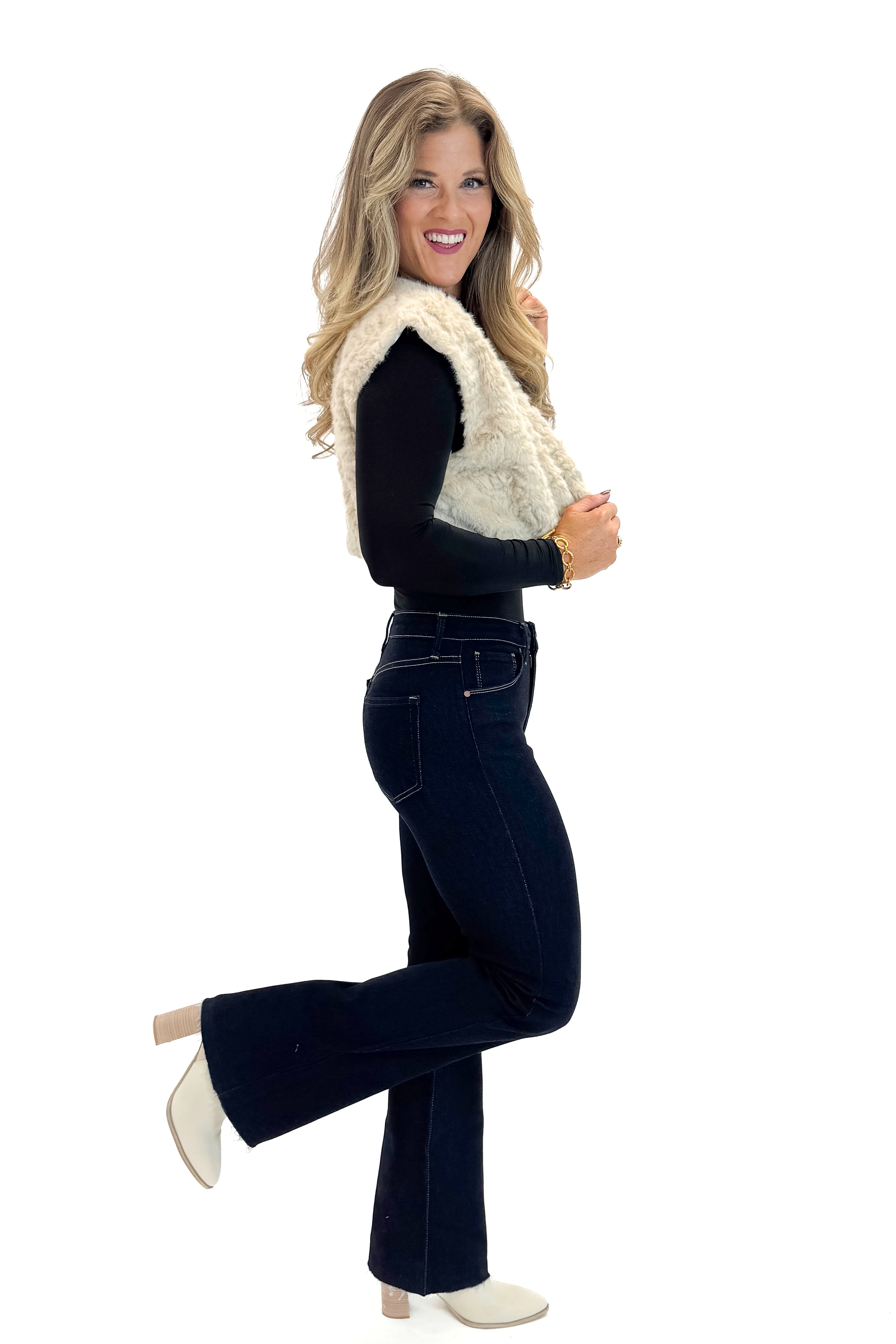 Side view of woman wearing Taupe Cropped Faux Fur Vest with jeans against white studio background.