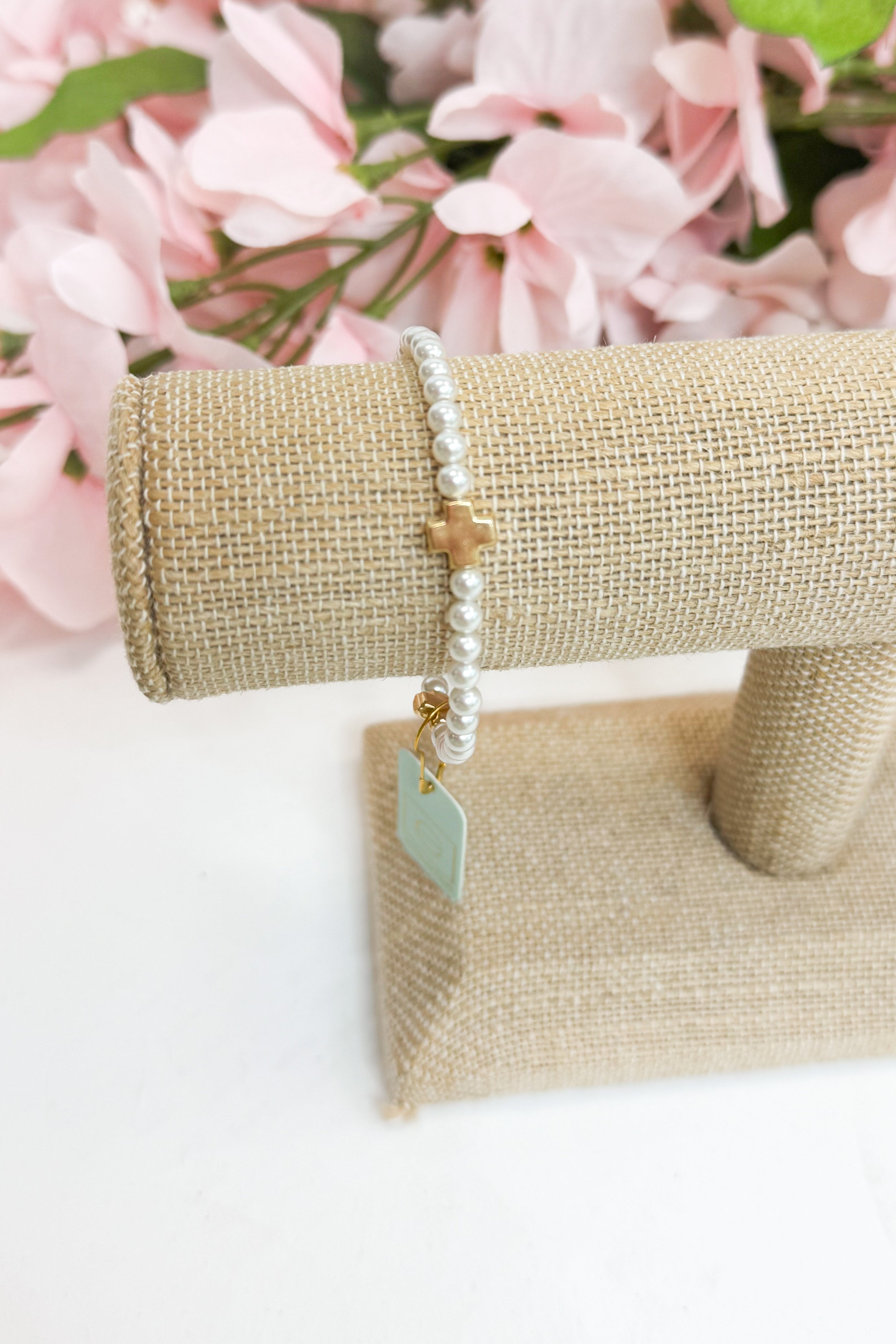 Pearl bracelet with a gold cross and tag on a beige jewelry stand with pink flowers in the background